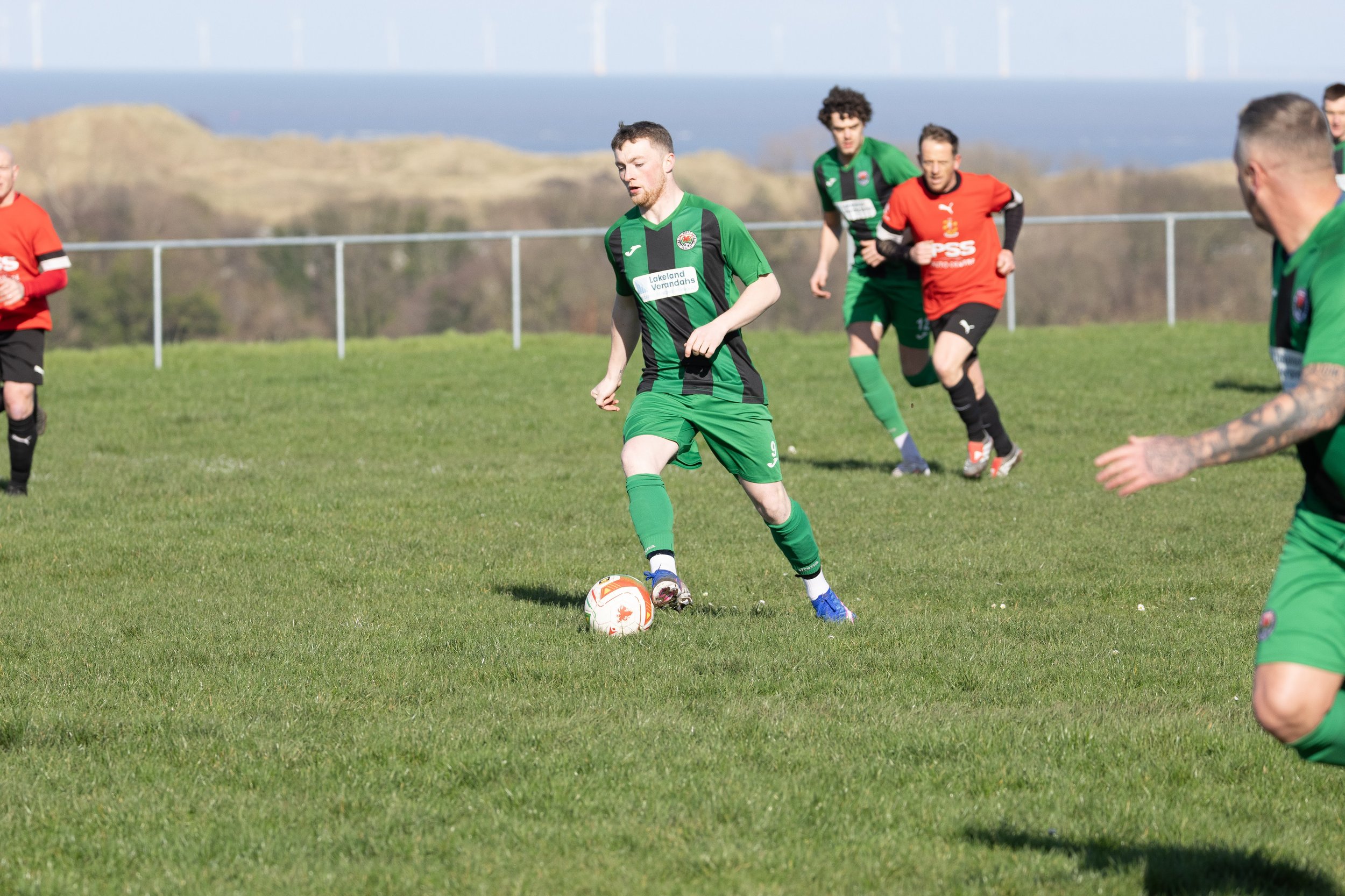 A soccer player in a green and black uniform controls the ball on a grassy field with other players and a scenic landscape with hills in the background.