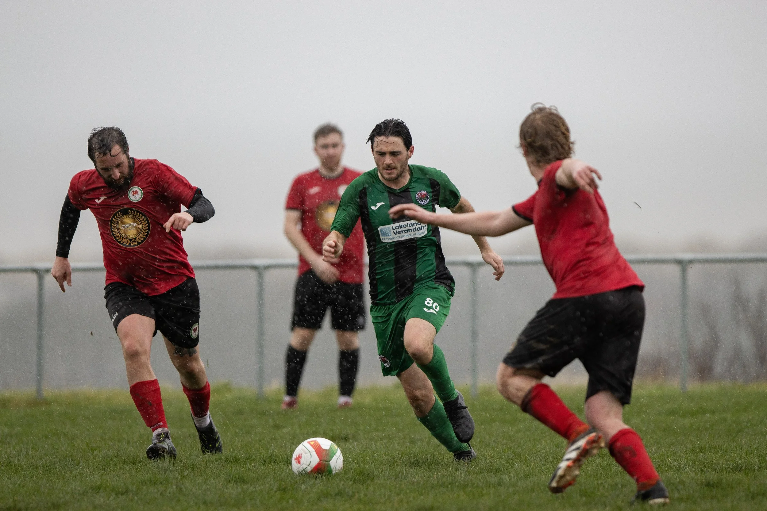 Soccer players competing on a wet field in rainy weather, with one player in green running towards the ball while opponents in red challenge him.