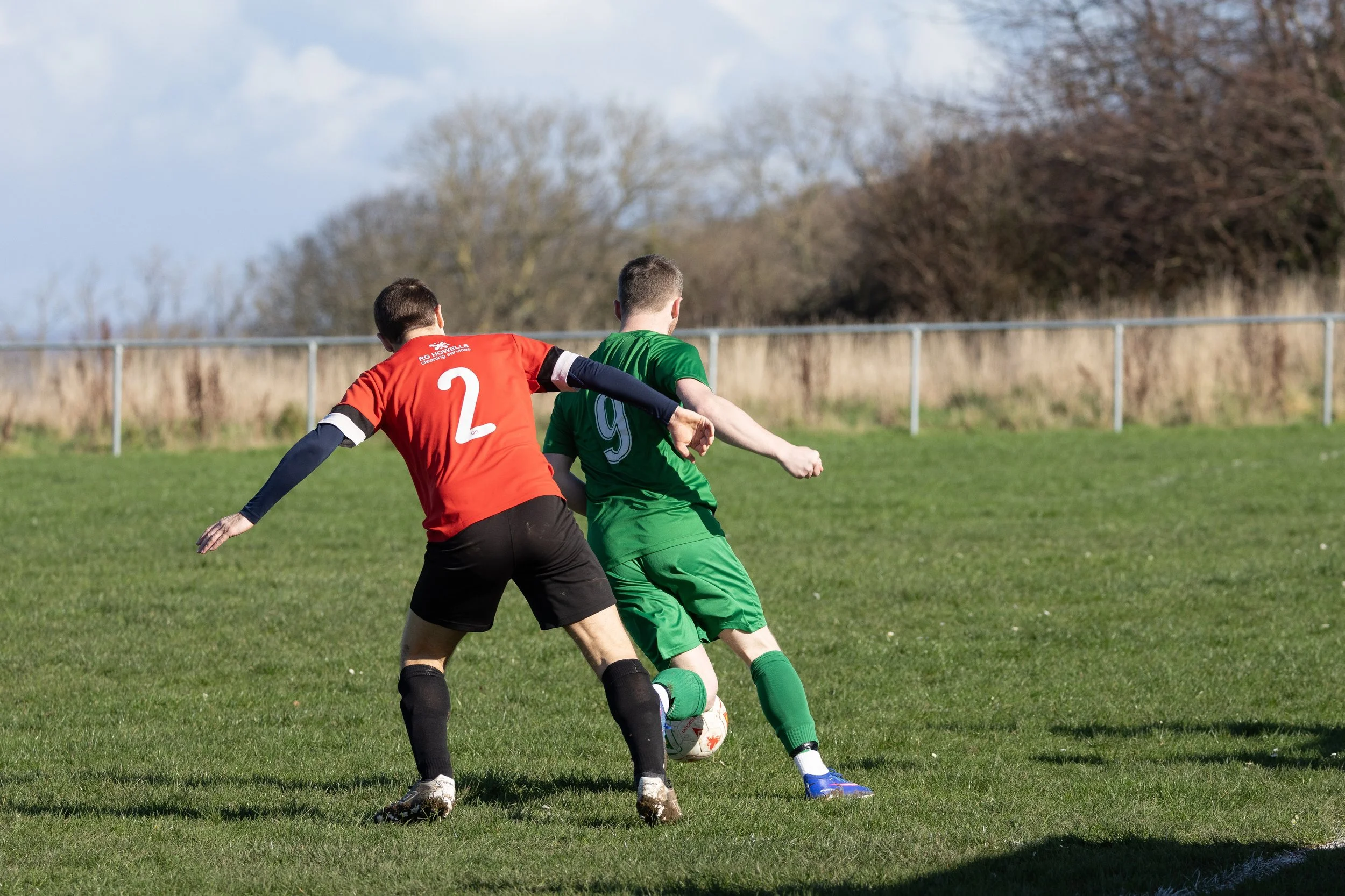 Two soccer players, one in a red jersey with the number 2 and the other in a green jersey with the number 9, competing for the ball on a grassy field.