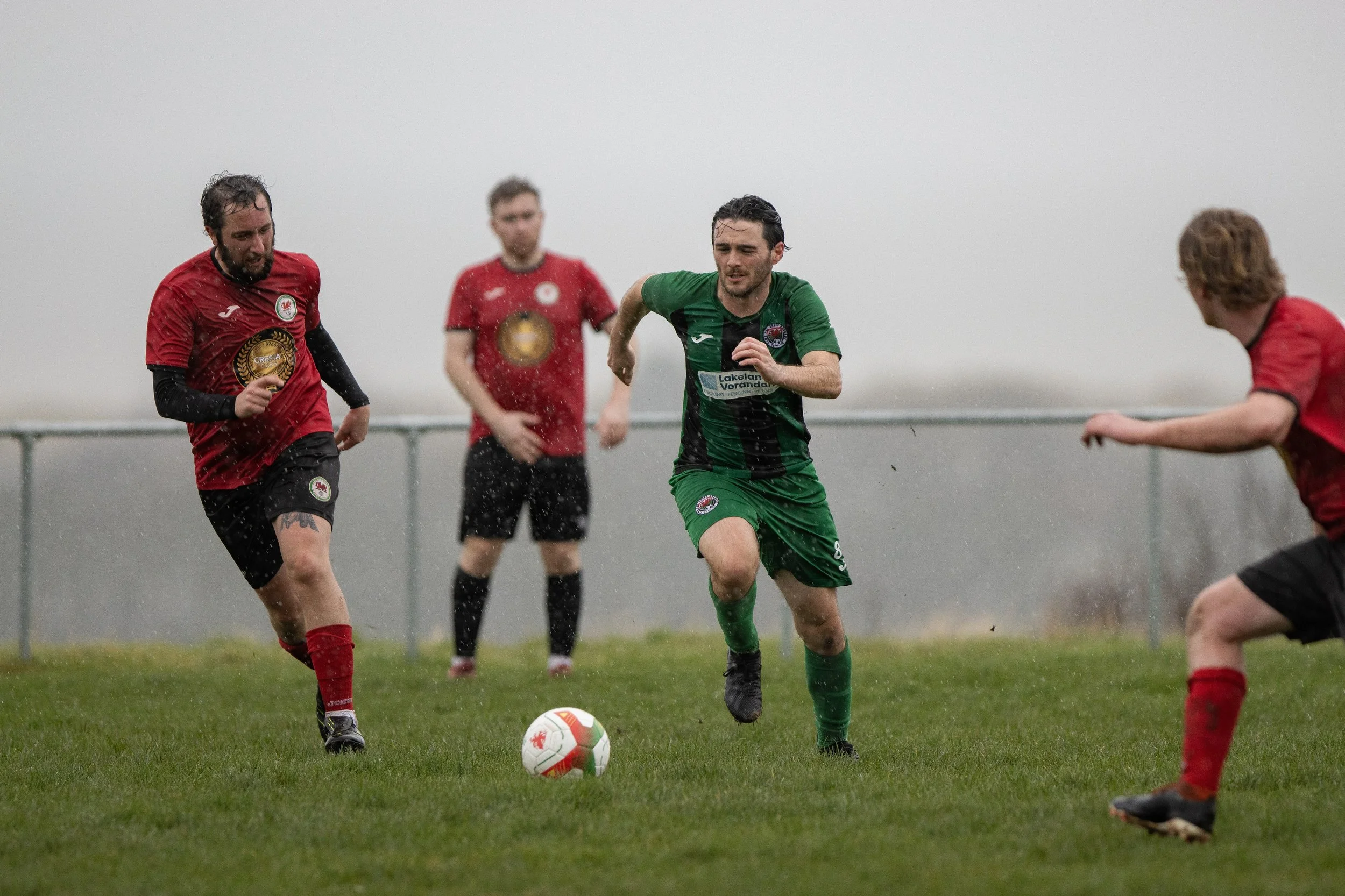 Four soccer players wearing red and green jerseys playing in rainy weather on a grass field, with one player in green kicking the ball.