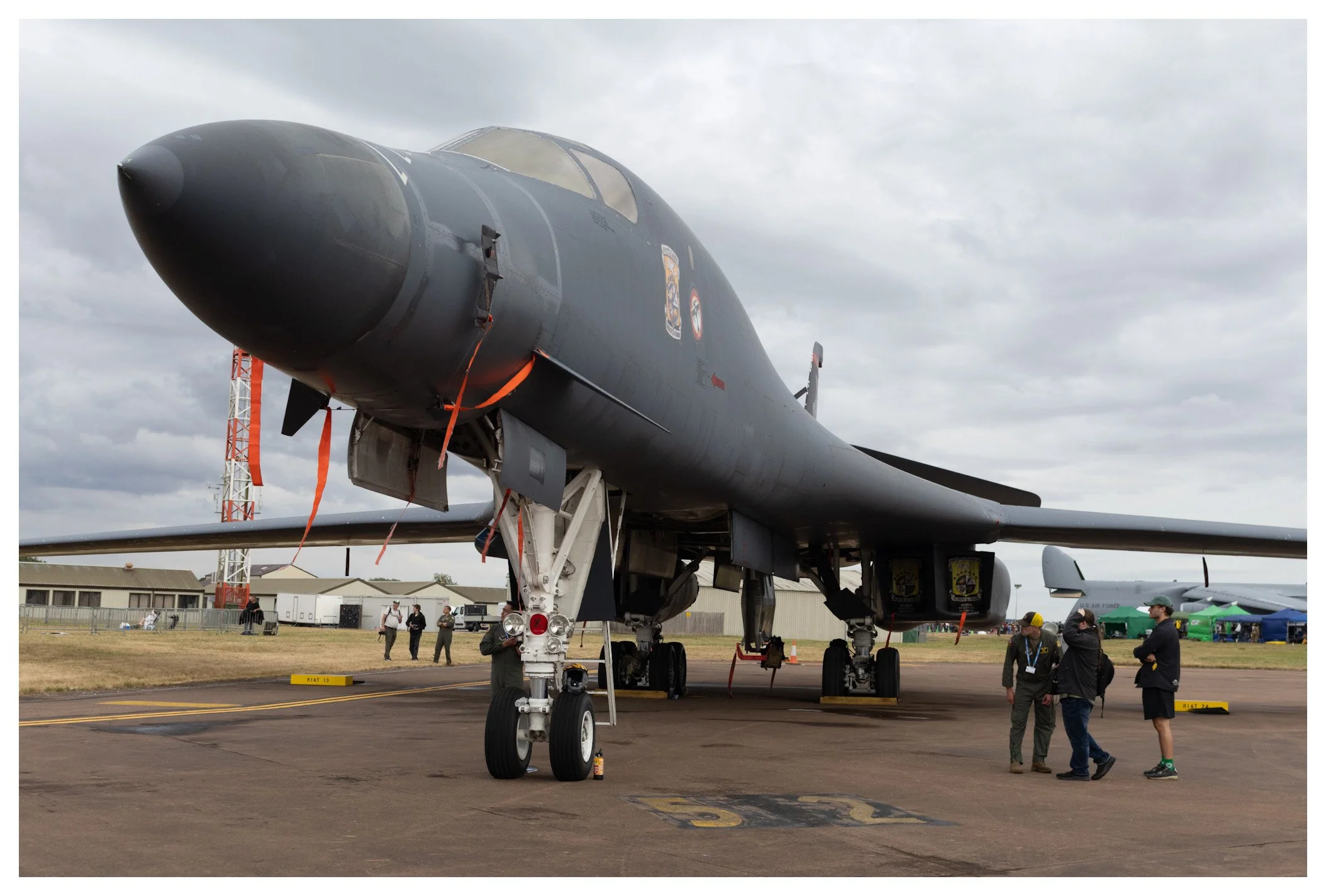 A large military jet fighter on display at an airfield, with people walking and observing around it under cloudy skies.
