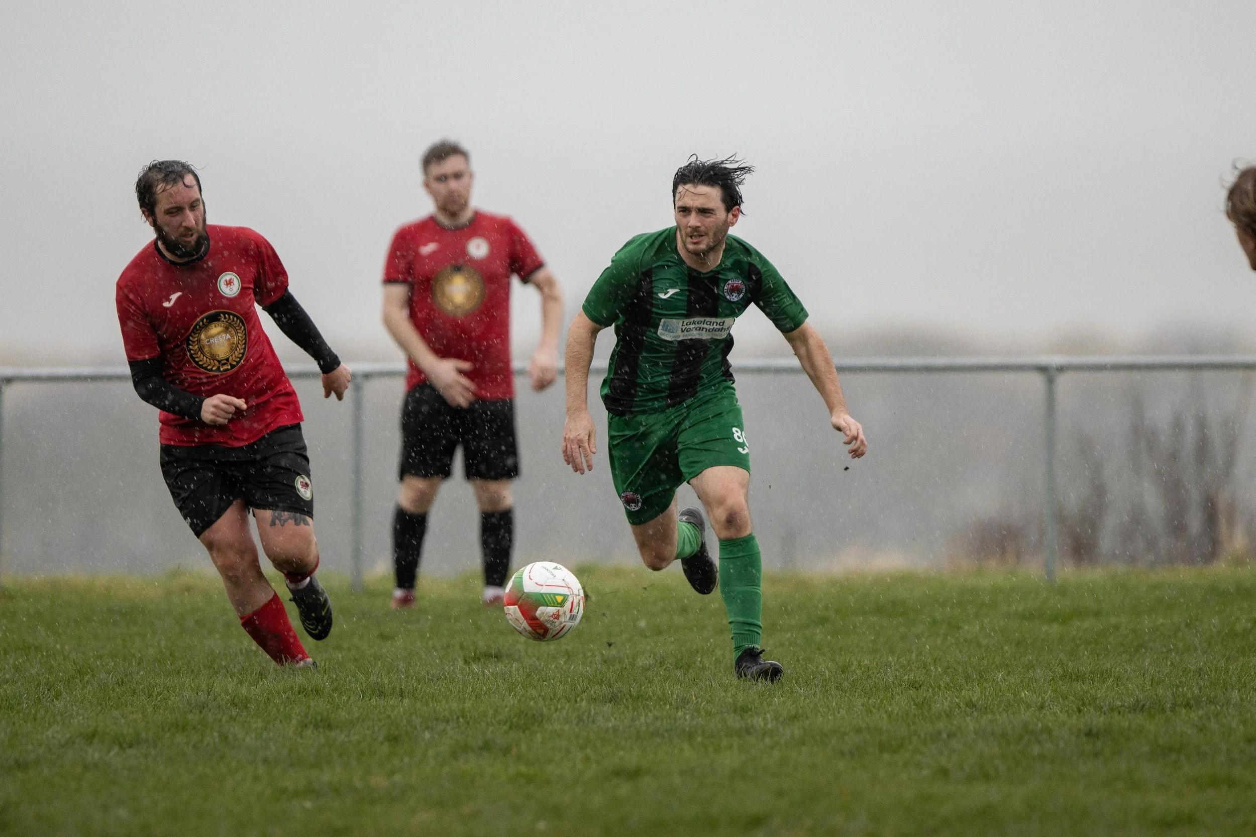 Soccer players sprint on wet grass field during rain, competing for the ball in a match.