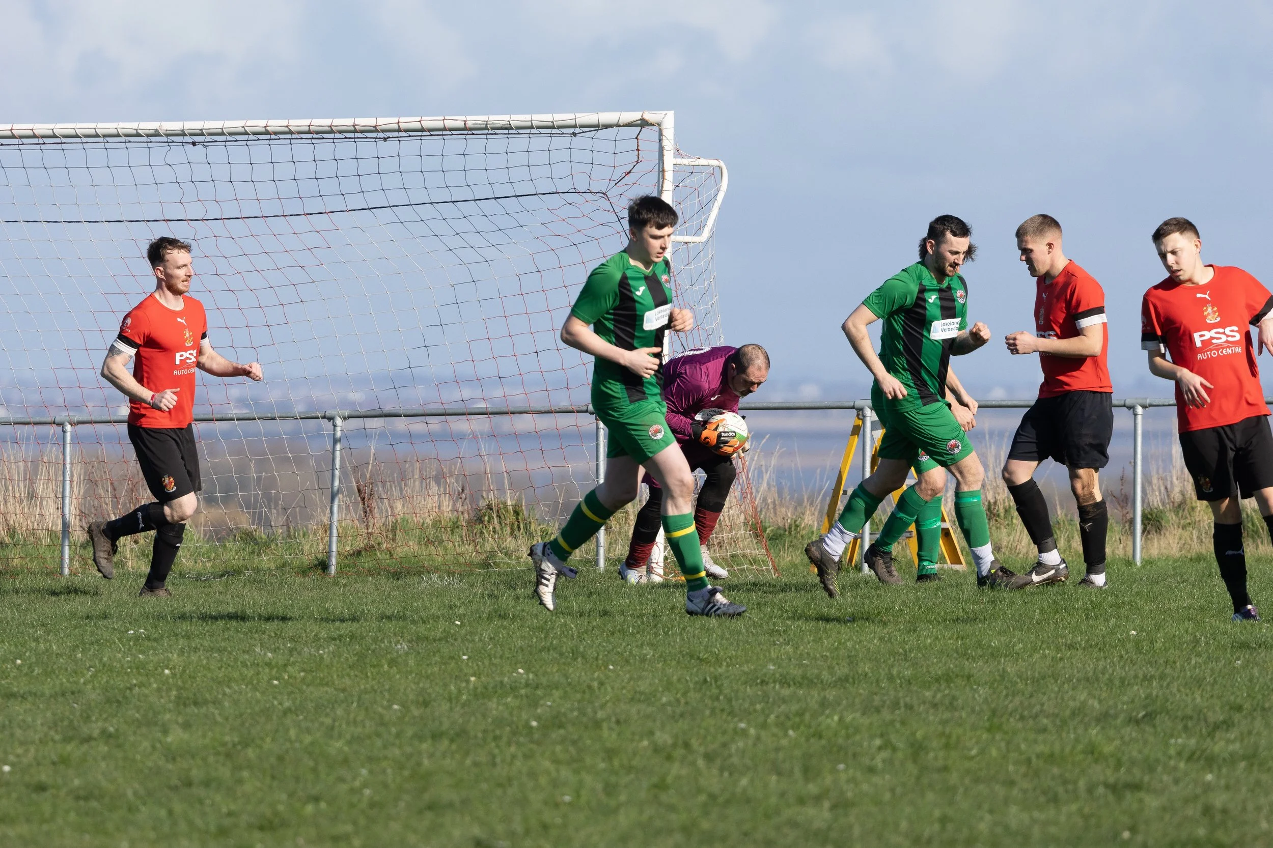 Soccer players near the goalpost, with one goalkeeper holding a ball and others standing nearby, on a grassy field under a partly cloudy sky.