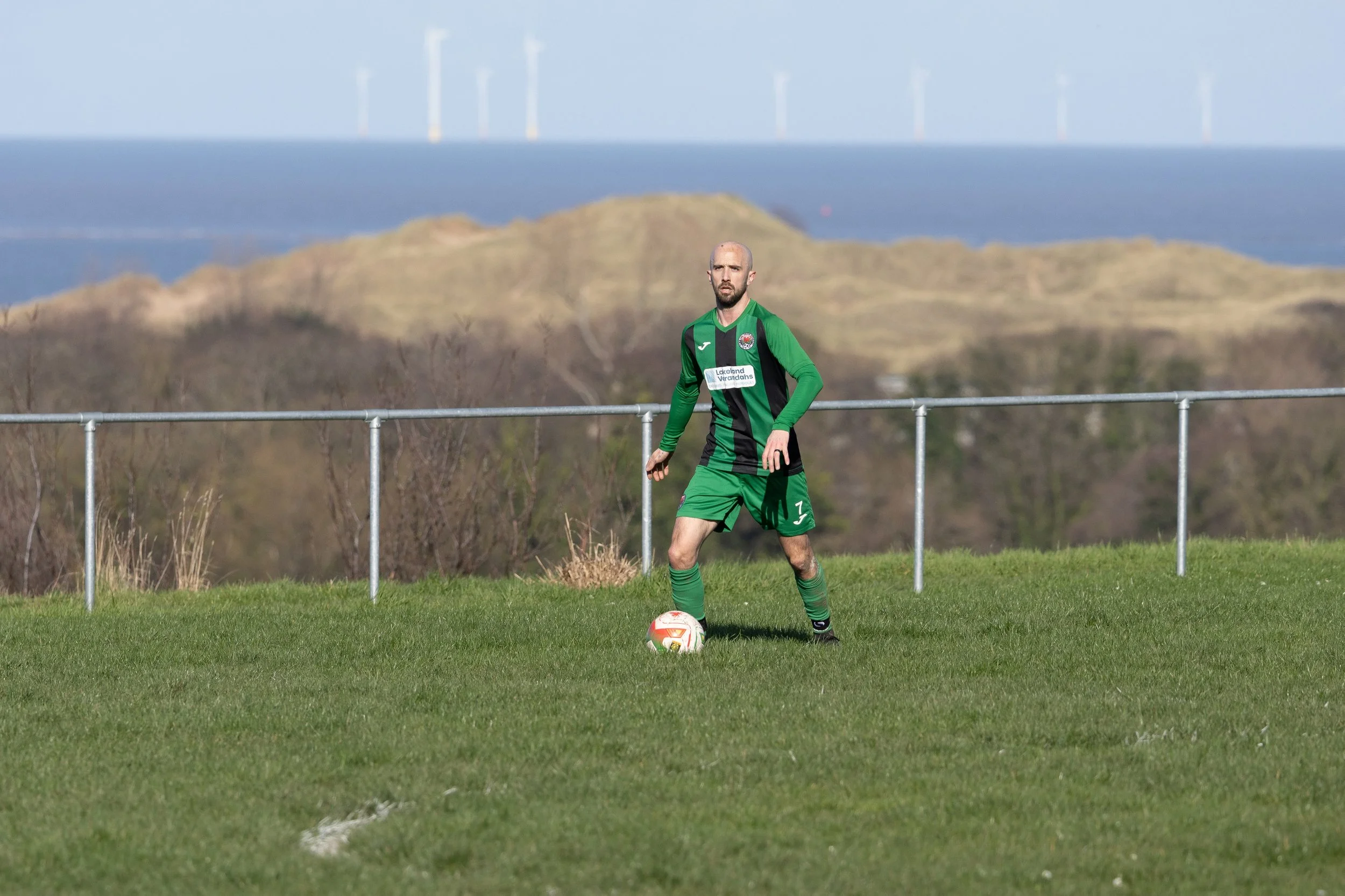 A male soccer player standing on a grassy field with a soccer ball at his feet, against a background of hills and wind turbines.