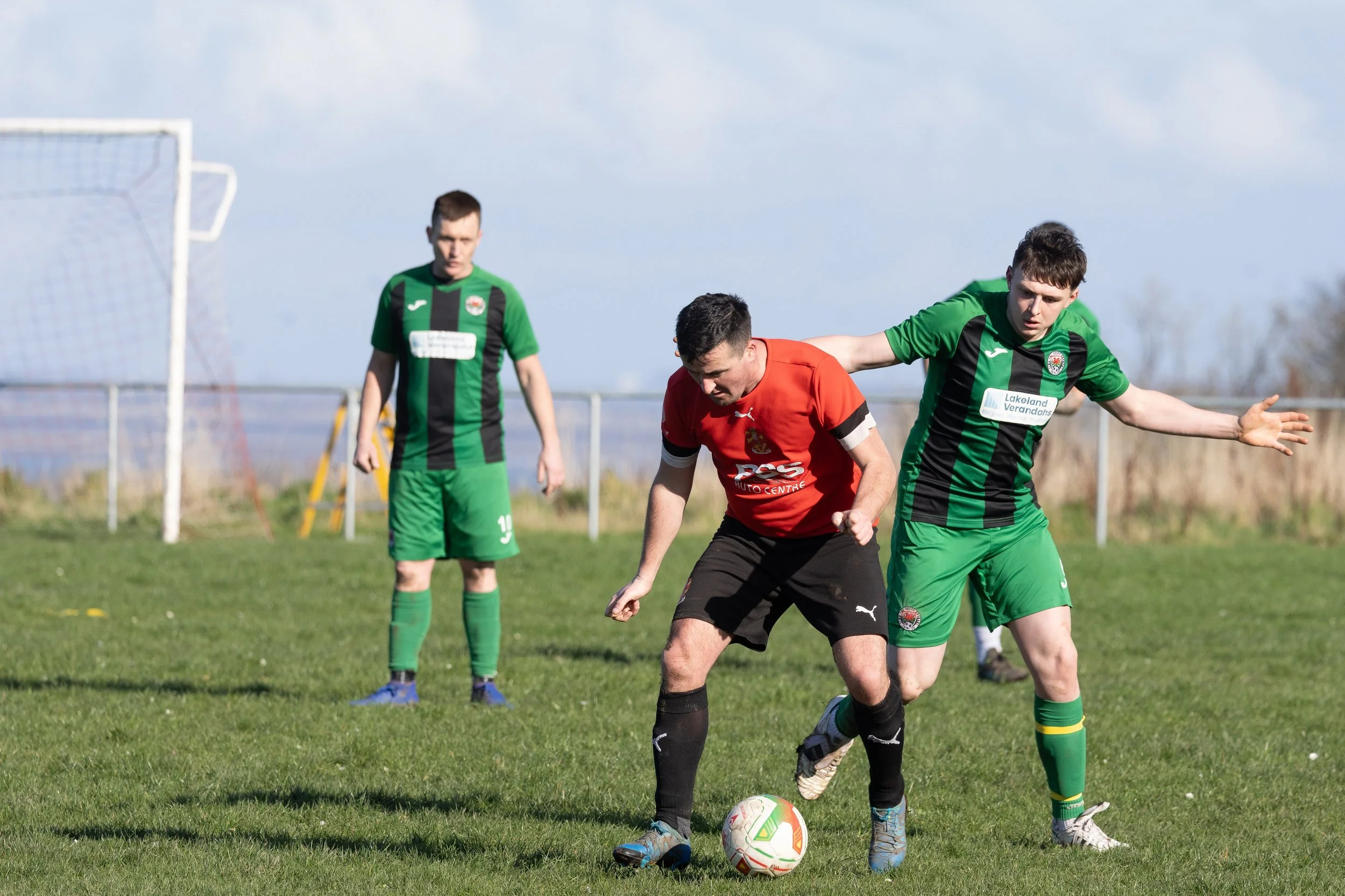 Three soccer players on a grassy field, one in a red jersey and two in green and black jerseys, competing for the ball with a goal in the background