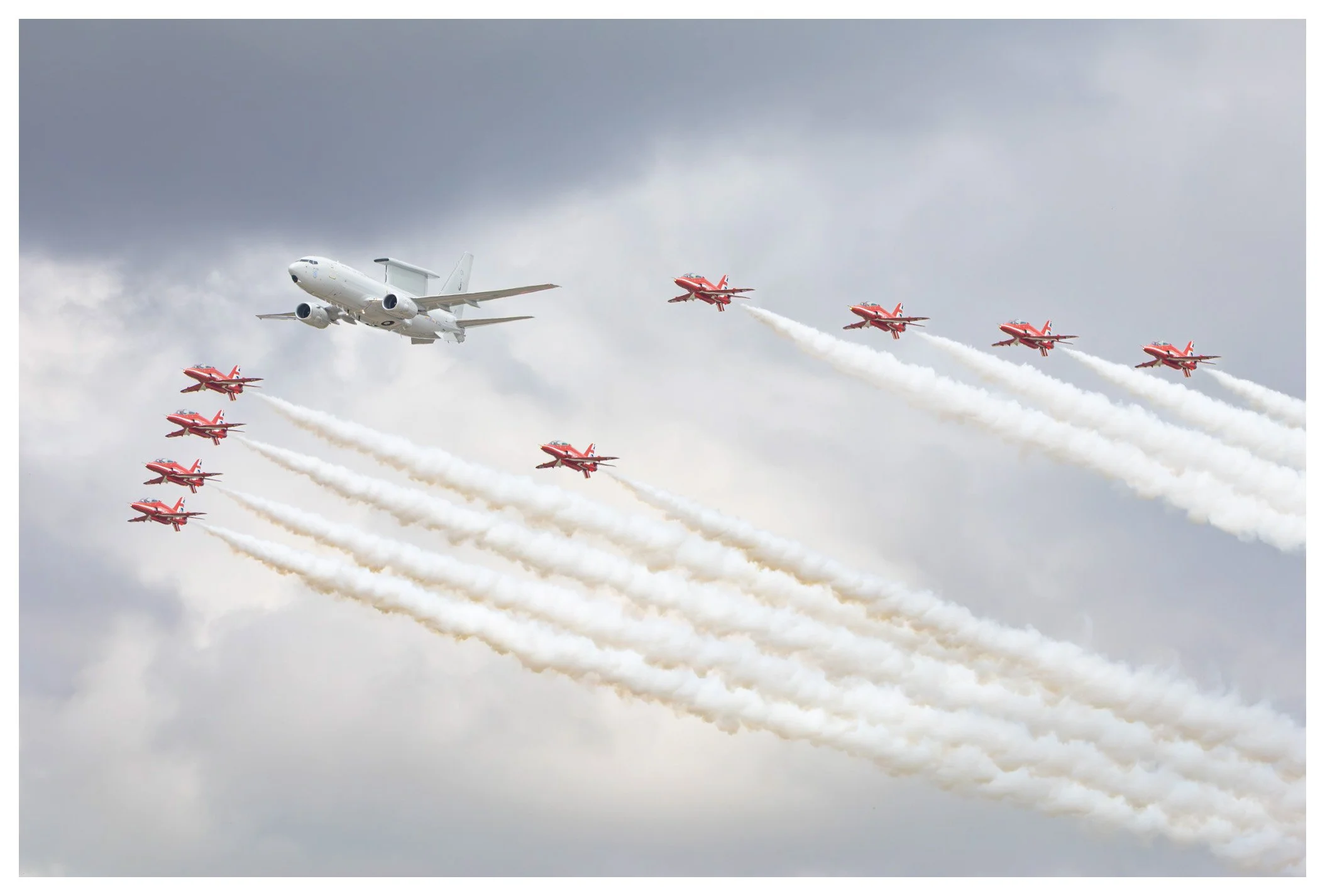 An air show with a large white passenger airplane flying alongside multiple red and white jet planes emitting white smoke trails against a cloudy sky.