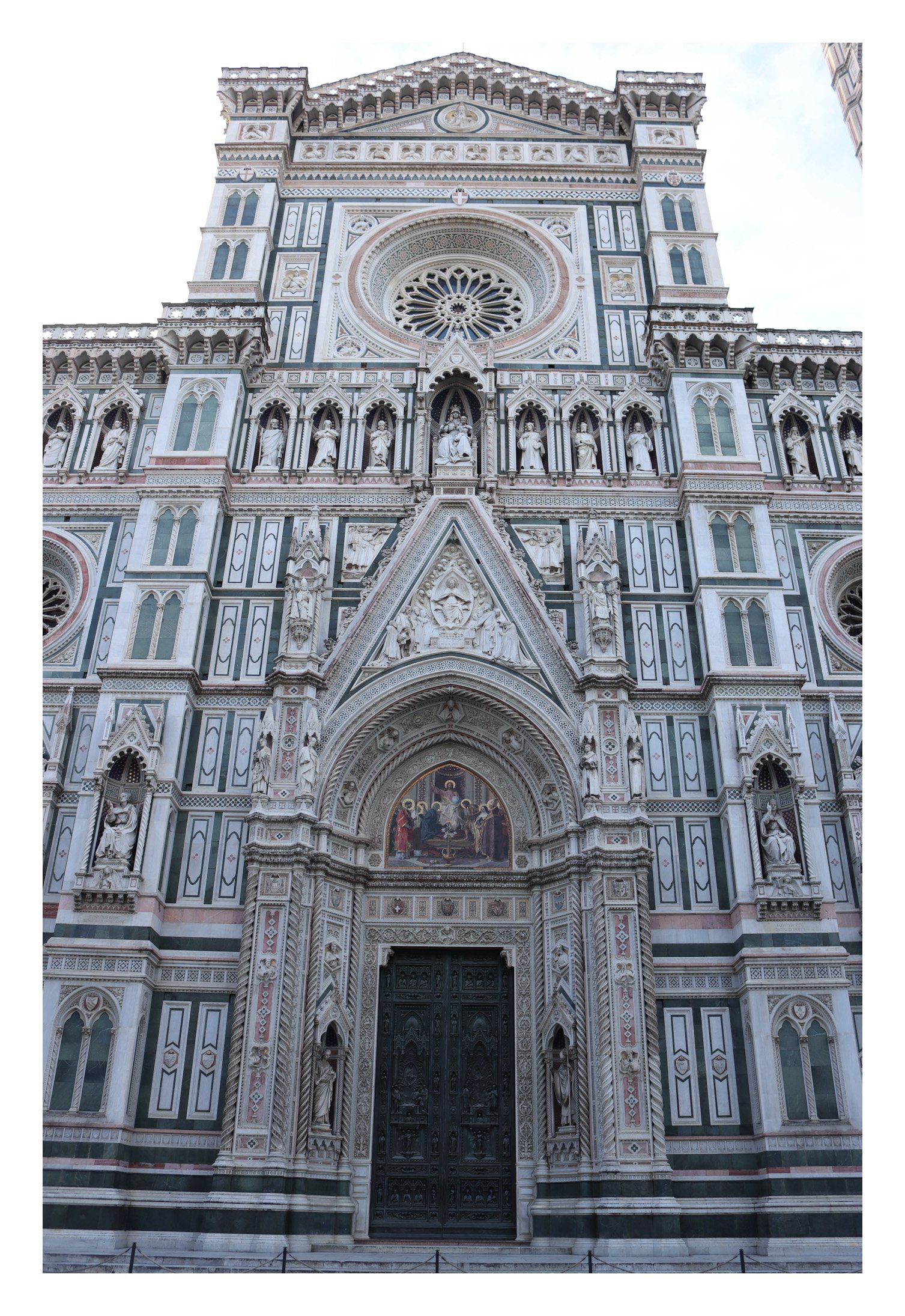 Front facade of the Florence Cathedral with intricate white, green, and pink marble details, statues, and a large circular stained-glass window.