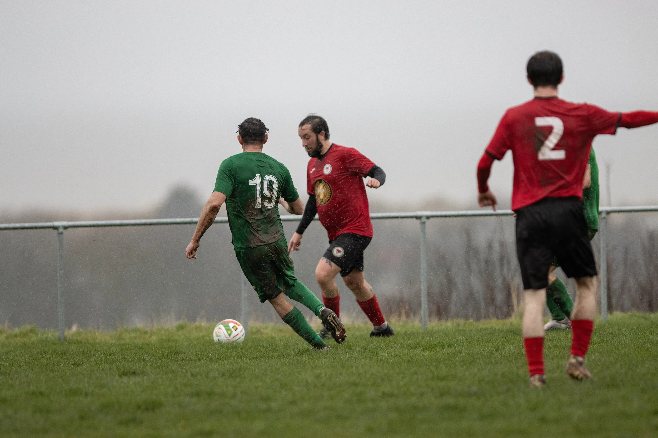 Soccer game in rain with players in green and red jerseys competing on a grassy field.