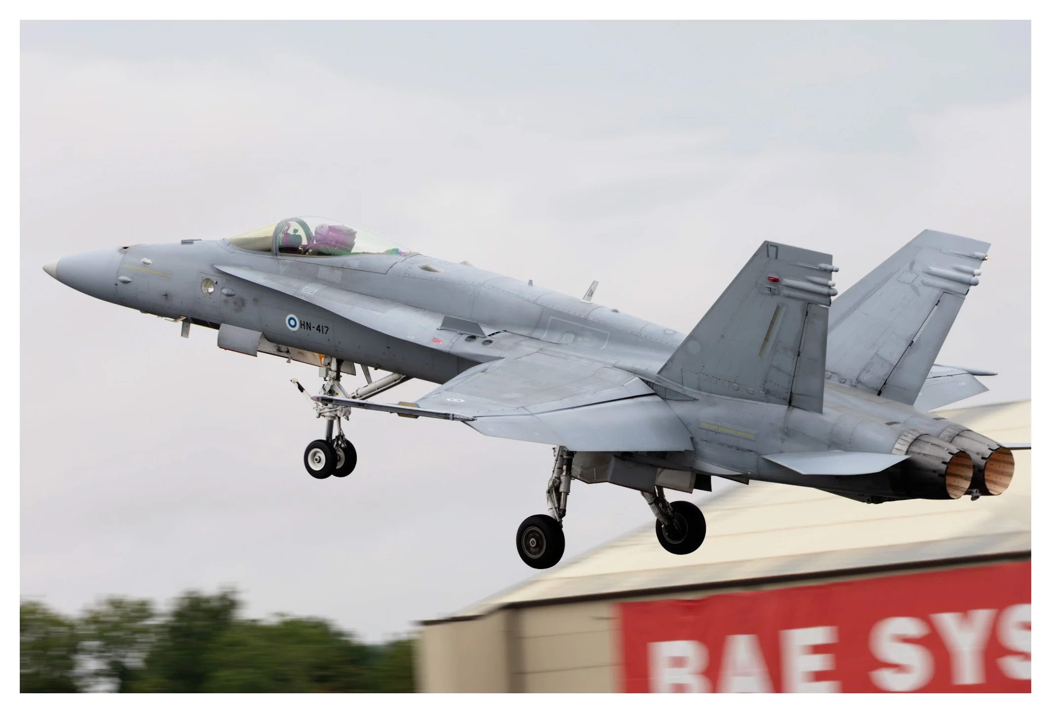 A gray fighter jet aircraft taking off, with landing gear extended, against a cloudy sky background and part of a red and white structure visible in the foreground.
