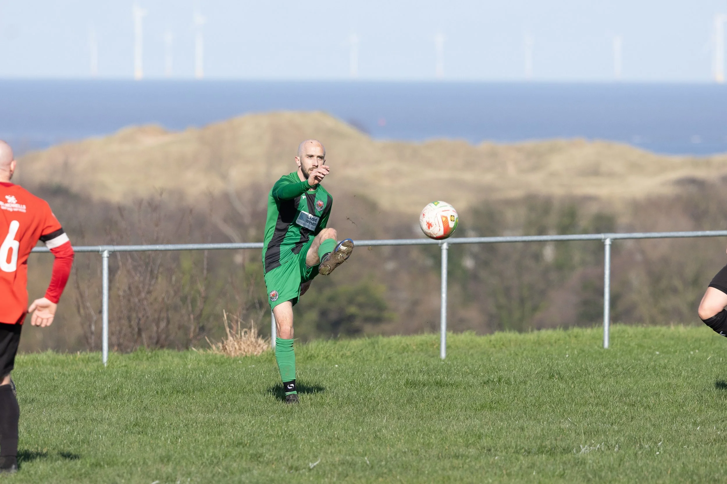 A soccer player in a green uniform practicing on the field, with a ball mid-air, and other players around, outdoors with hills and wind turbines in the background.