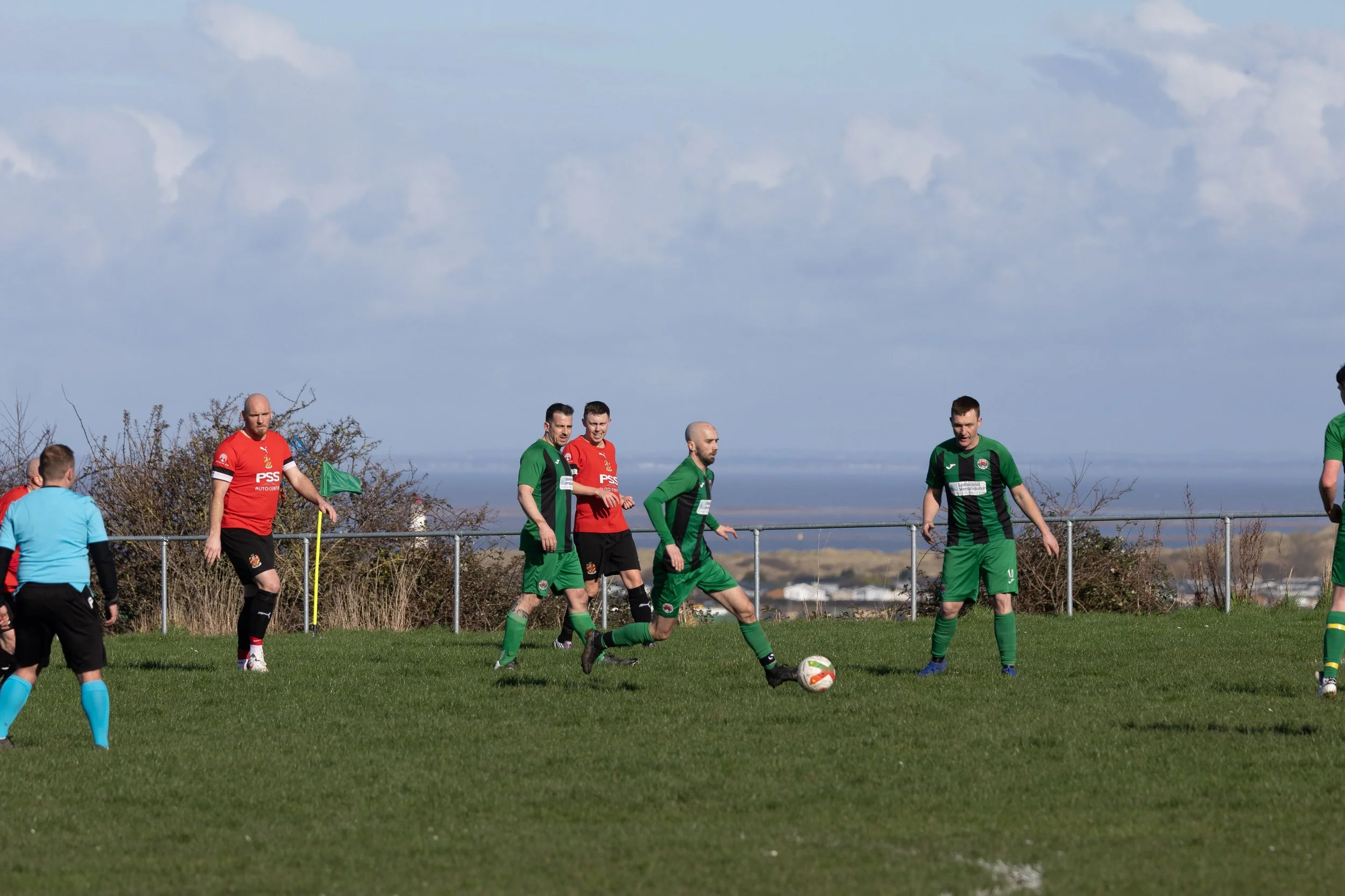 Soccer players on the field during a match, with some wearing red and others green jerseys, a referee in blue, and a cloudy sky in the background.