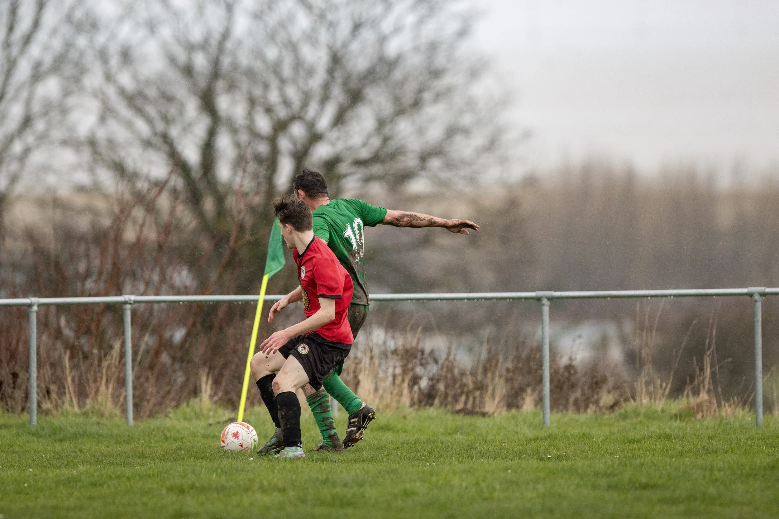 Two boys playing soccer outdoors on a grassy field, one in a red jersey and the other in a green jersey, with trees and a cloudy sky in the background.
