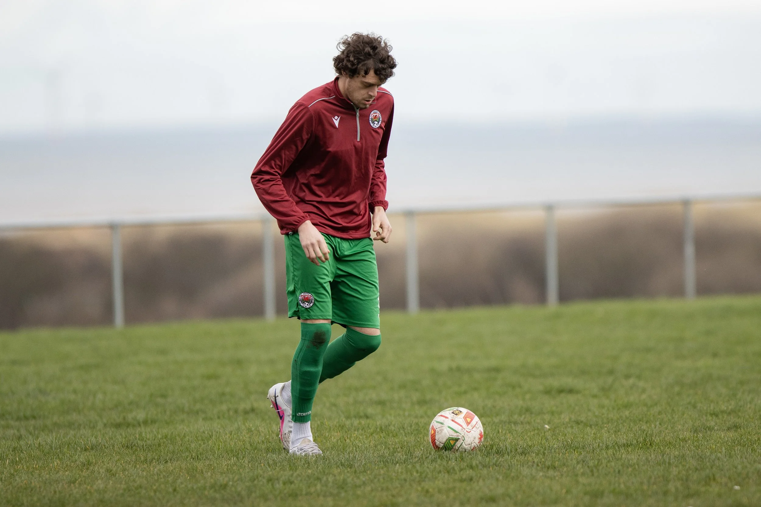 A soccer player wearing a maroon and green uniform standing on a grassy field, looking down at a soccer ball.
