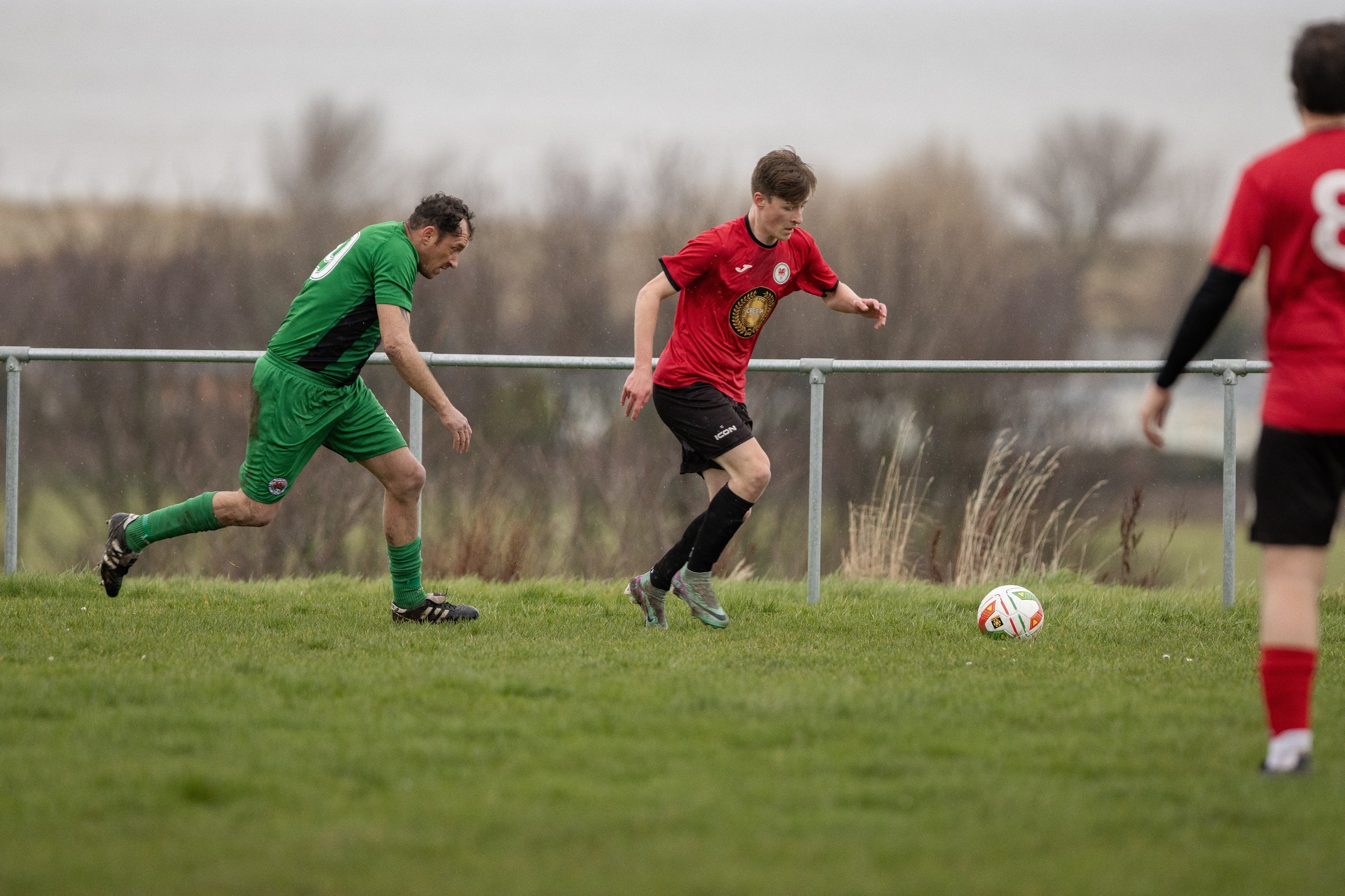 Soccer players competing on a grassy field during daytime, one in a green uniform and two in red uniforms, near a metal railing in a rural setting.