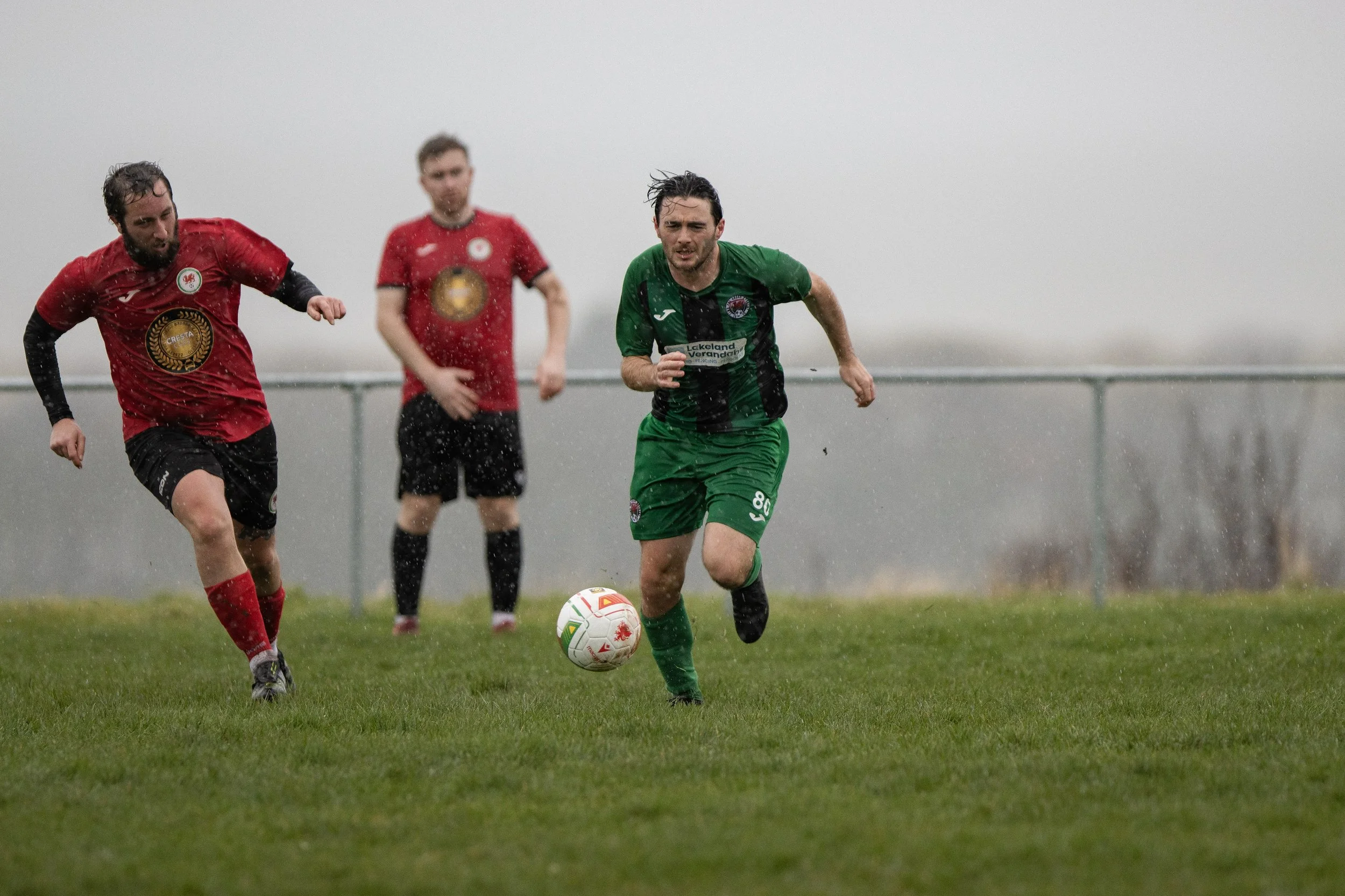 Three soccer players competing in the rain during a match on a grassy field. Two players wear red jerseys and black shorts, while the third player wears a green and black jersey with green shorts. The player in green is running towards the ball in fr