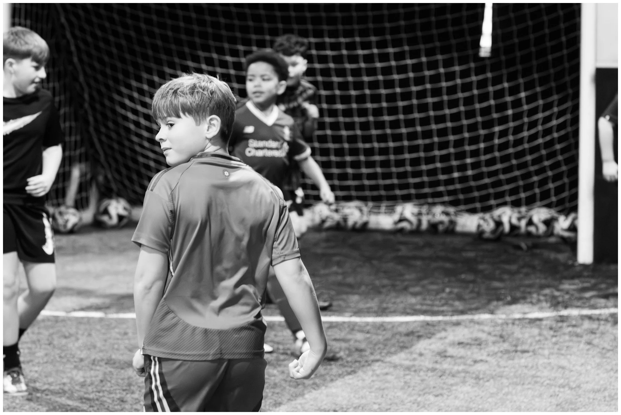 Children playing soccer on an indoor field with a net in the background, with one boy in the foreground making a fist gesture.