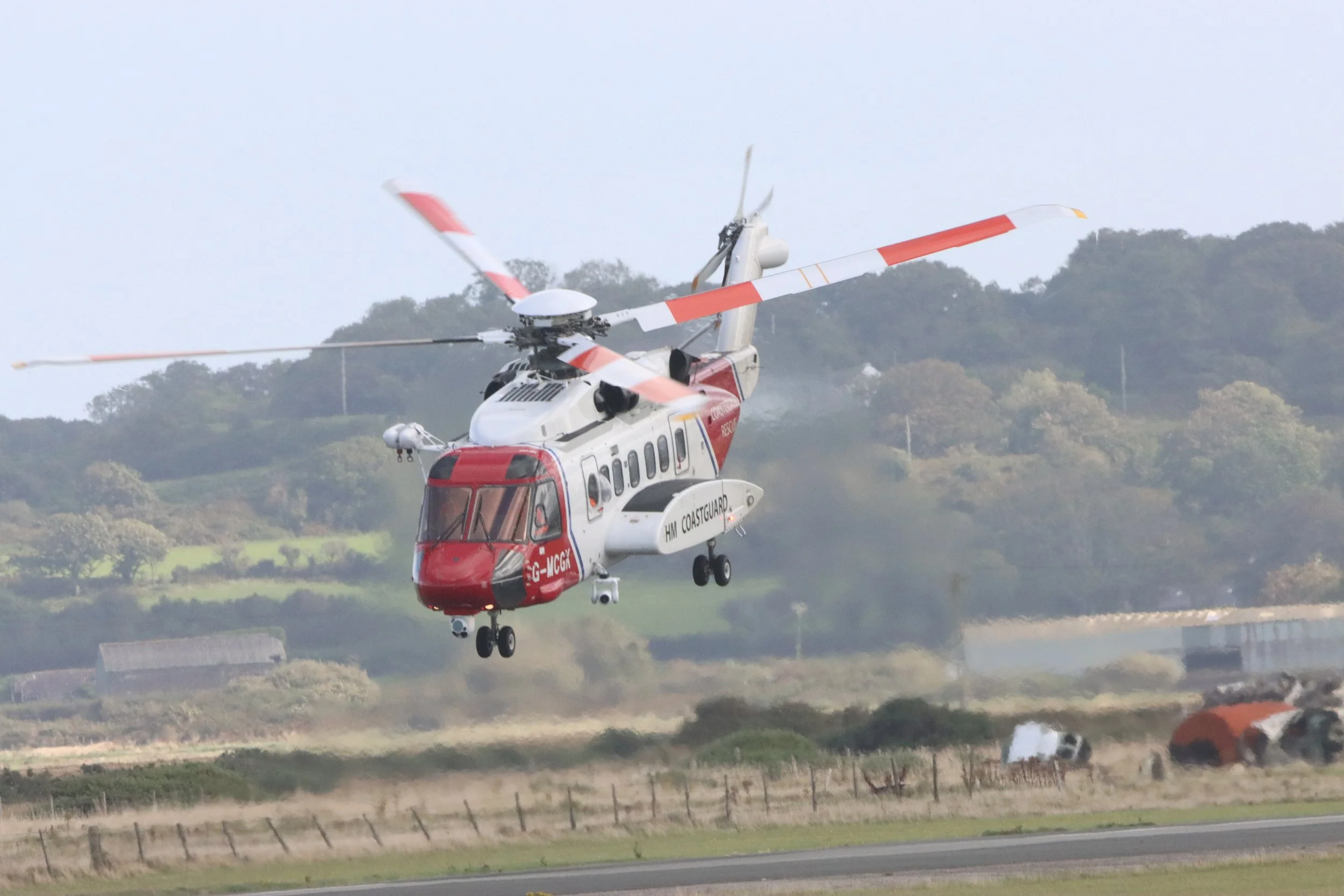 A Coastguard helicopter in flight above the ground, with hills and trees in the background. The helicopter is white and red, with landing gear extended and spinning rotor blades.