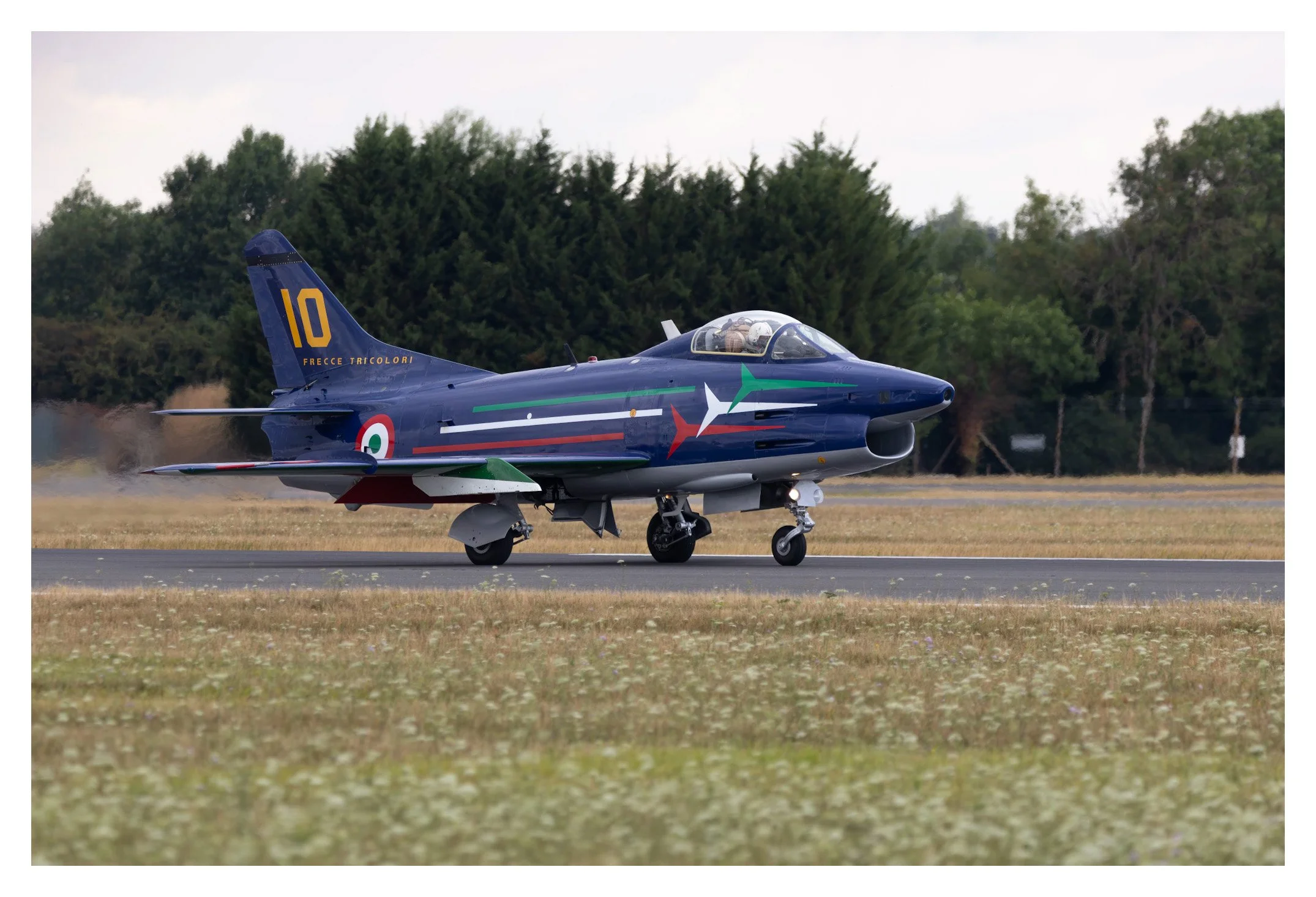Italian Air Force fighter jet on runway, blue with green, white, and red stripe, with the number 10 and Italian markings, taking off amid dust and grass, with trees in background.