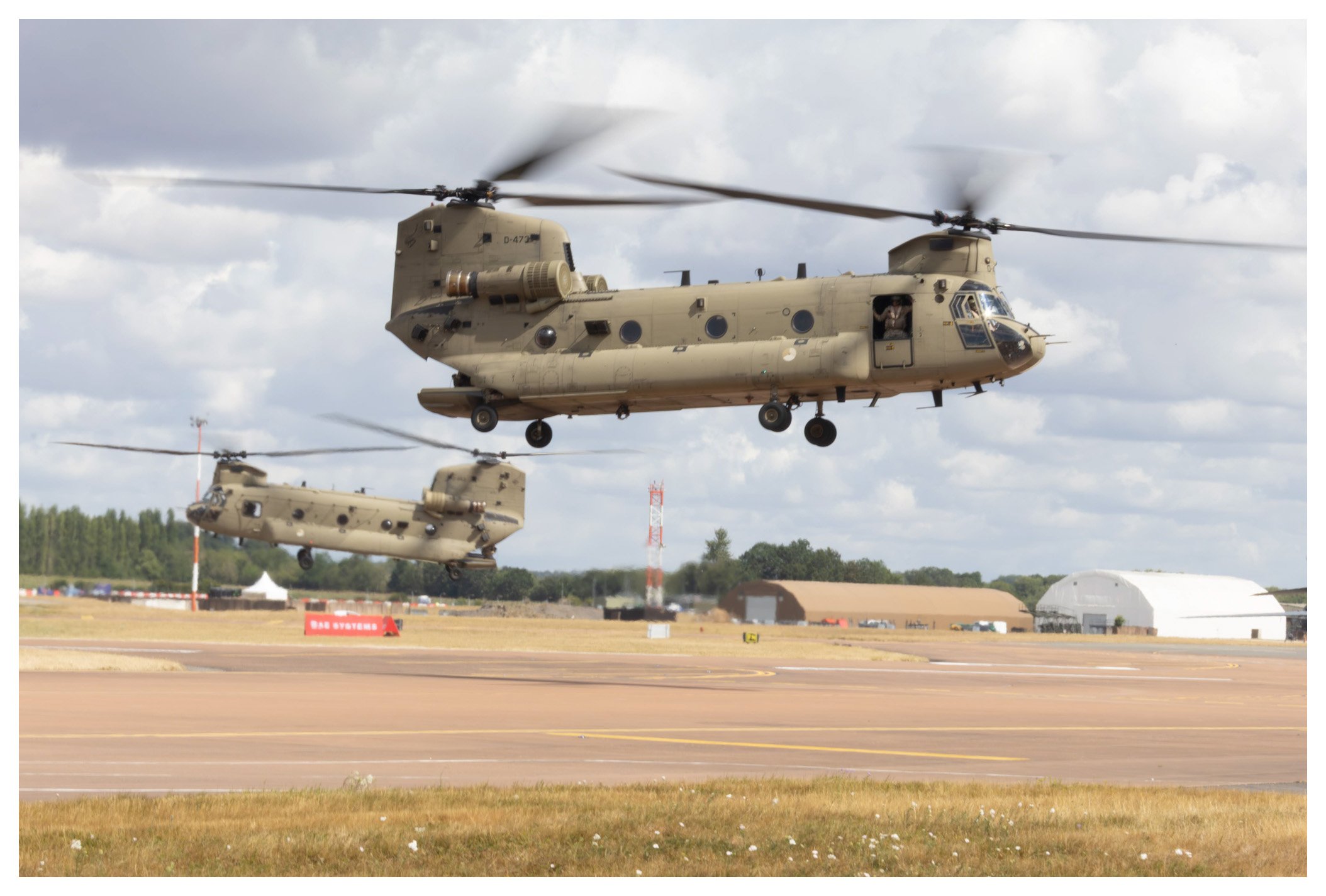 Two military helicopters flying low over an airfield during daytime, with hangars and a cloudy sky in the background.