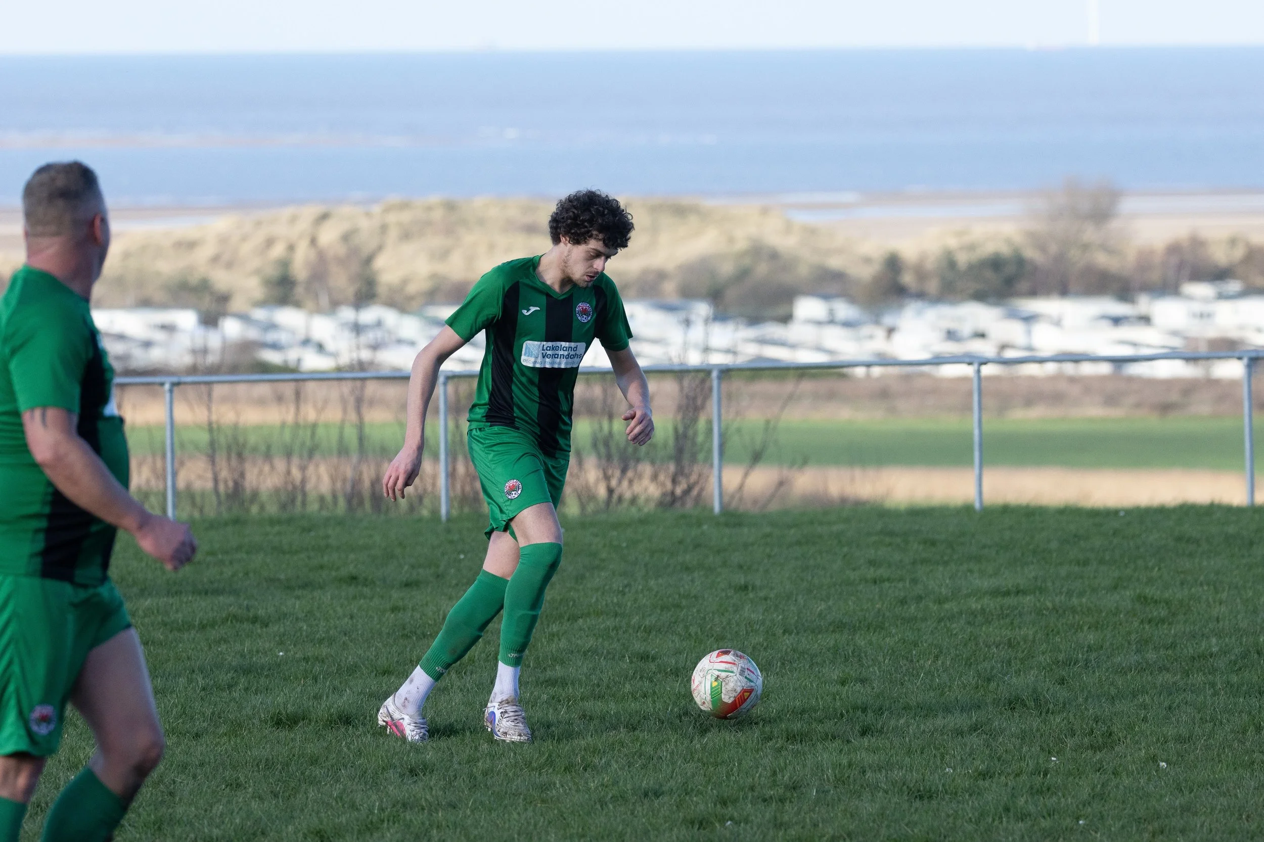 A soccer player in a green uniform about to kick a soccer ball on a grassy field, with another player partially visible nearby and a scenic background featuring water and distant land.