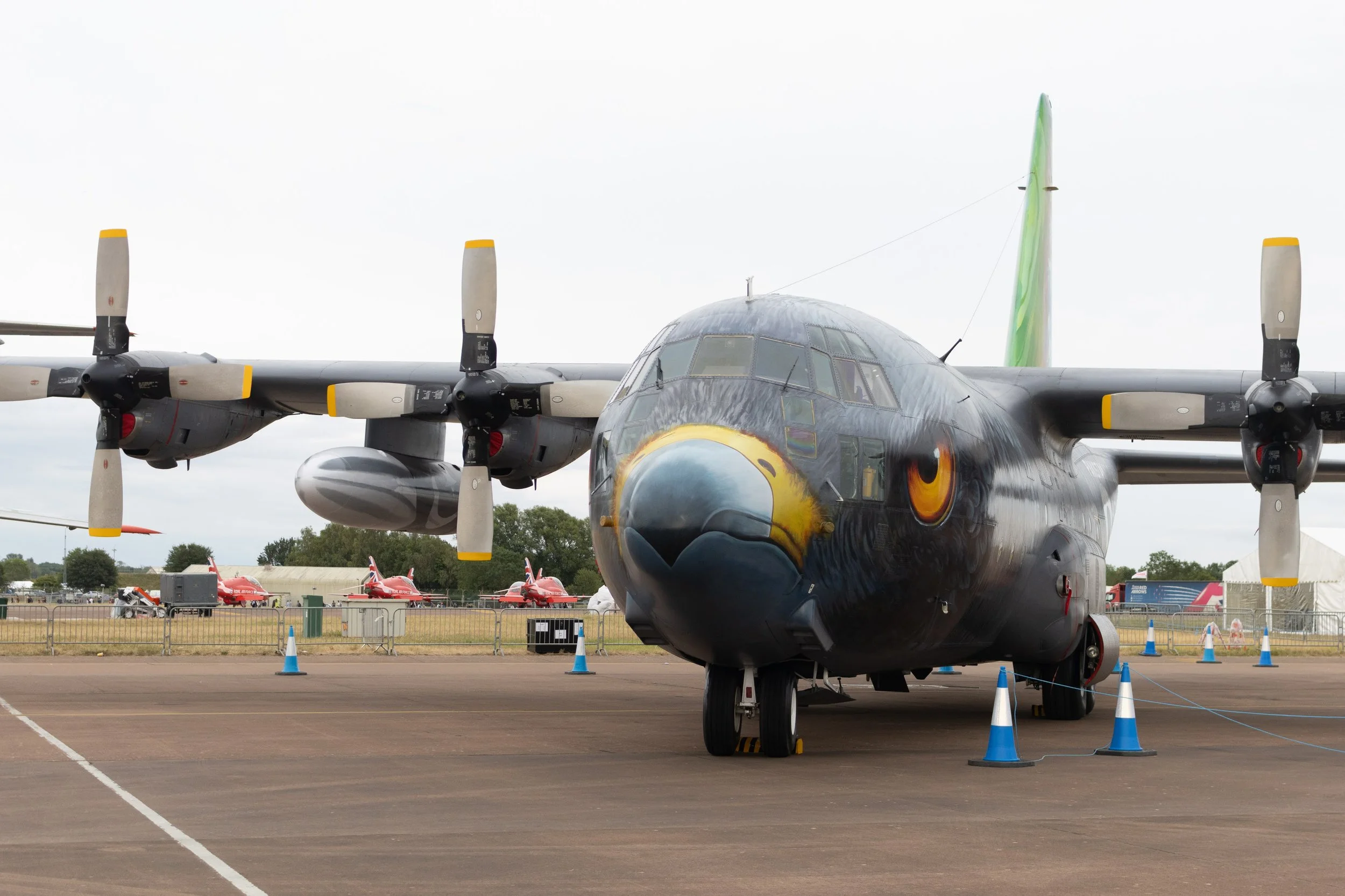 A propeller airplane painted with an eagle face and eyes on the front, parked on a tarmac at an airshow, with several red aircraft models and tents in the background.