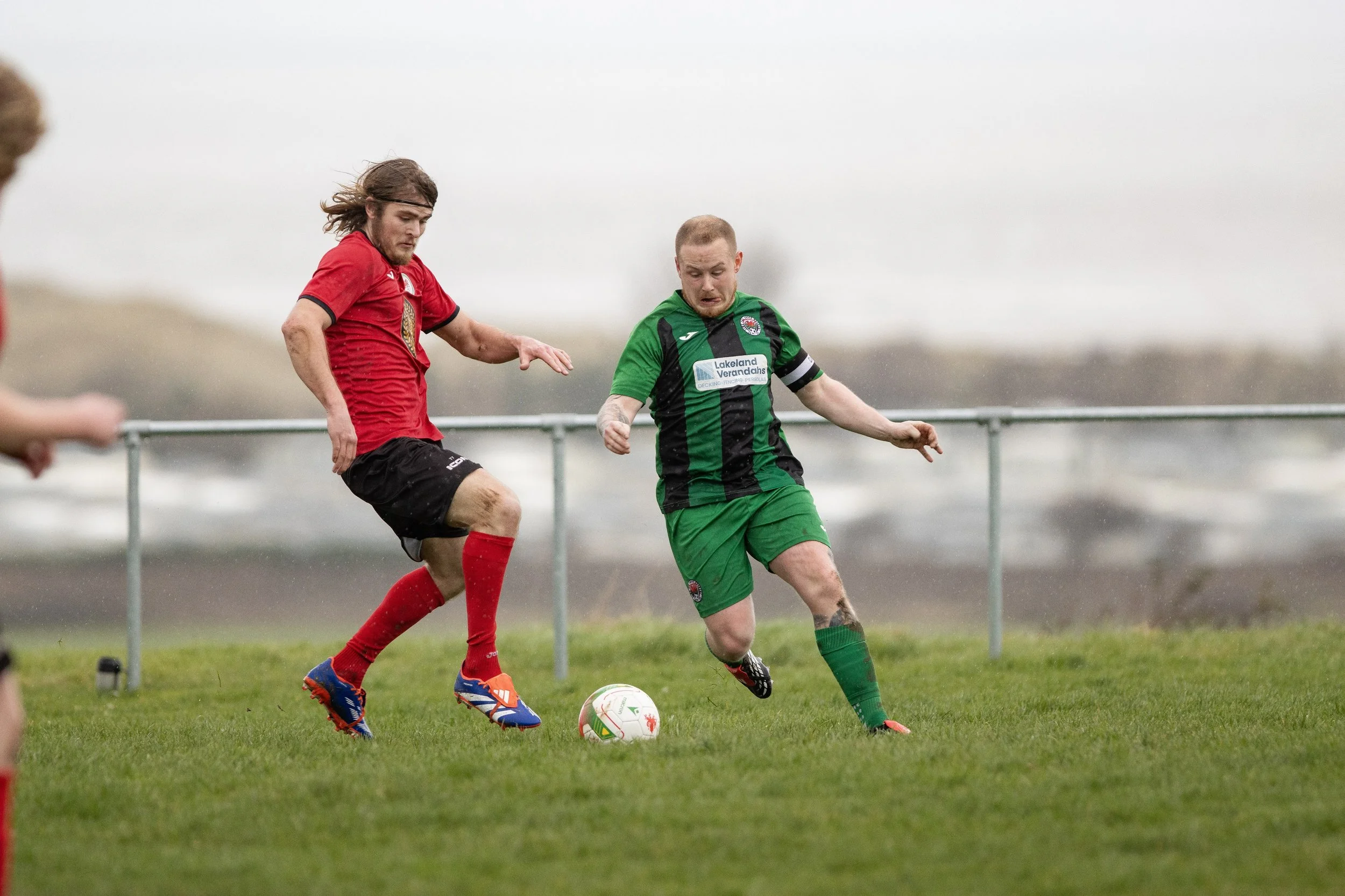 Two soccer players competing for the ball on a grass field, with a background of cloudy sky and distant ocean.