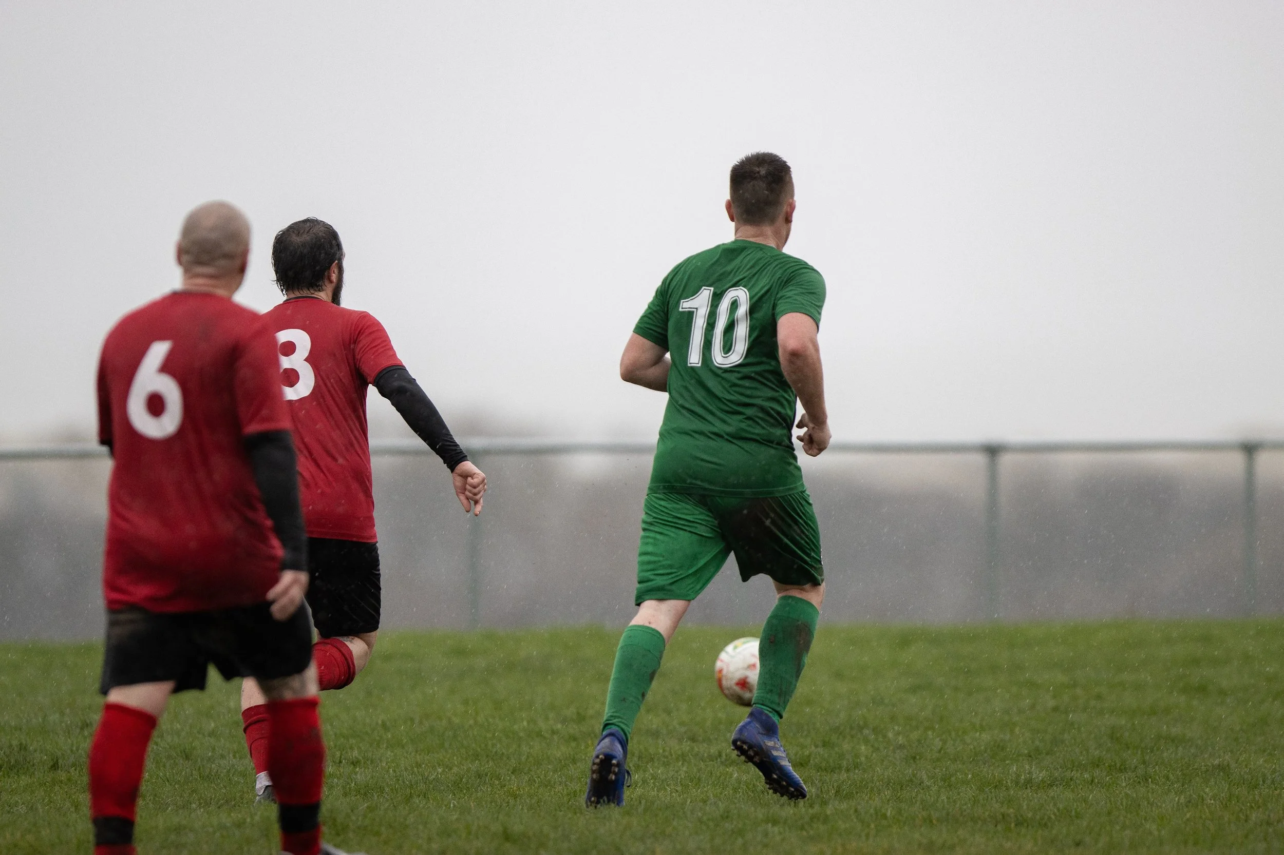 A soccer player in a green uniform with the number 10 on his back runs towards the ball on a rainy day, with two defenders in red jerseys nearby on a grassy field.