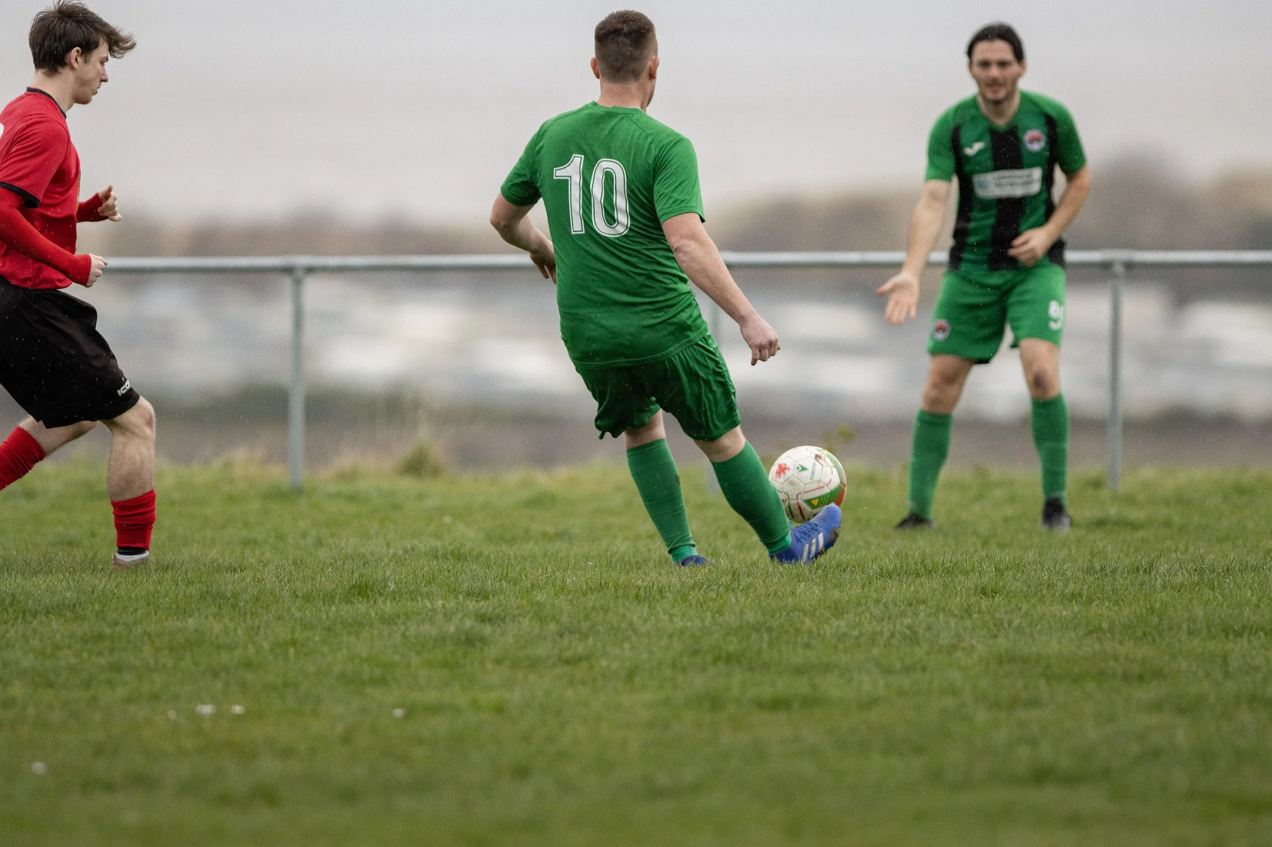 Three men playing soccer on a grassy field, with two wearing green jerseys and one in red, during an overcast day.