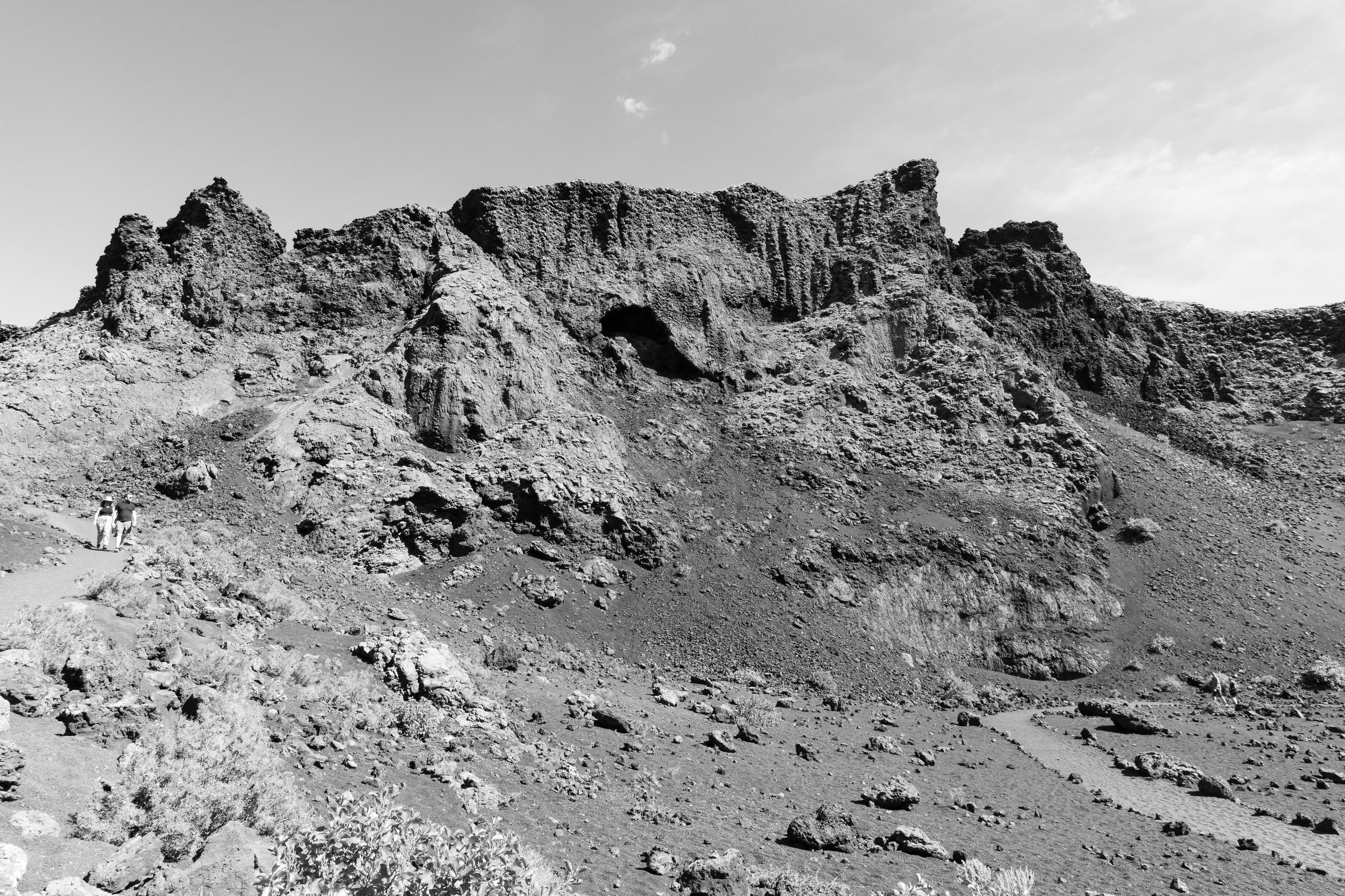 A black and white photograph of a rocky mountain landscape with steep slopes and a natural arch in the middle. Two people are walking along a trail in the lower left corner.