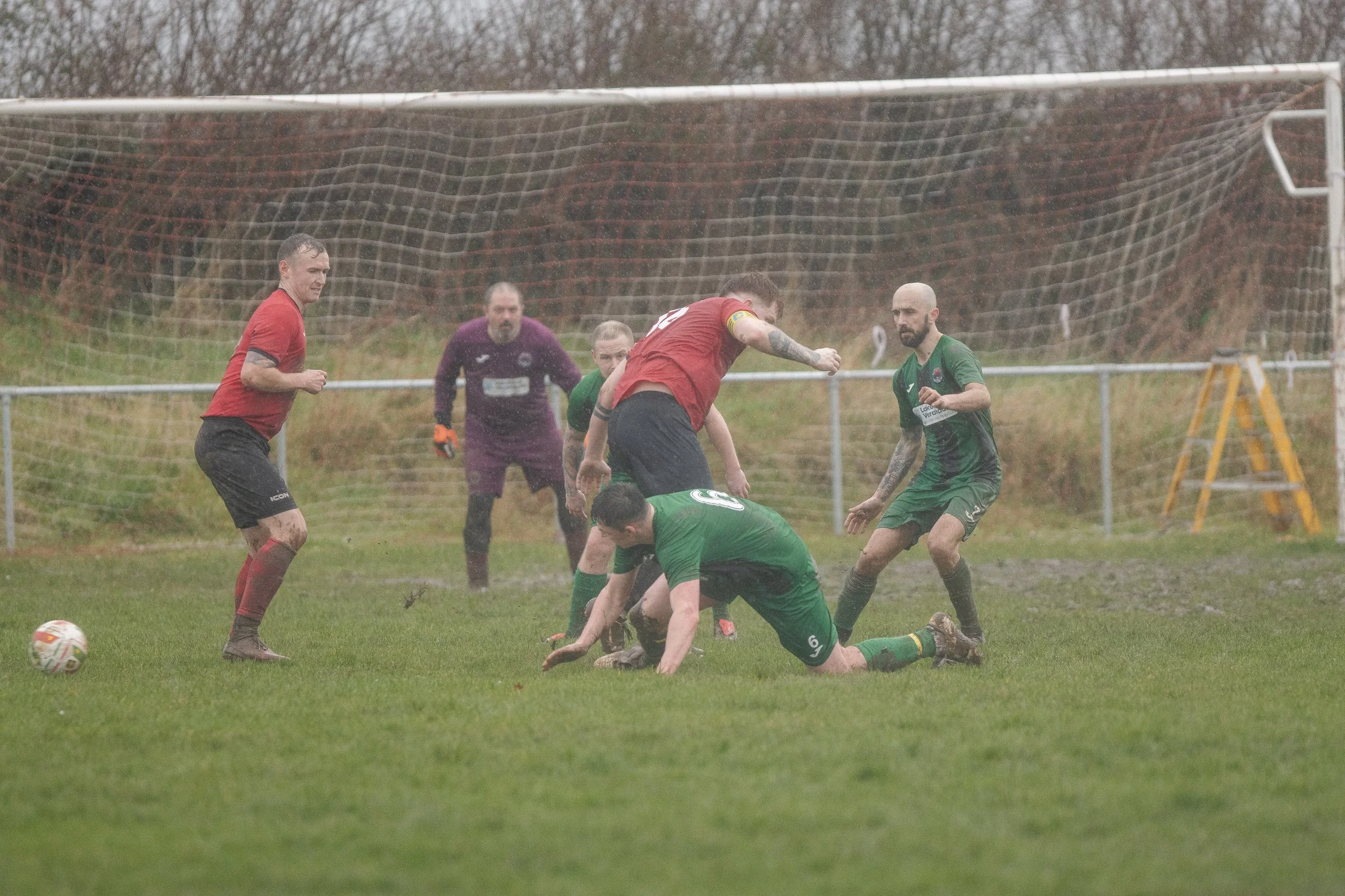Soccer players competing for the ball on a muddy field during a match, with one player in a red jersey jumping over players in green jerseys, and a goalkeeper standing in the background near the goal.