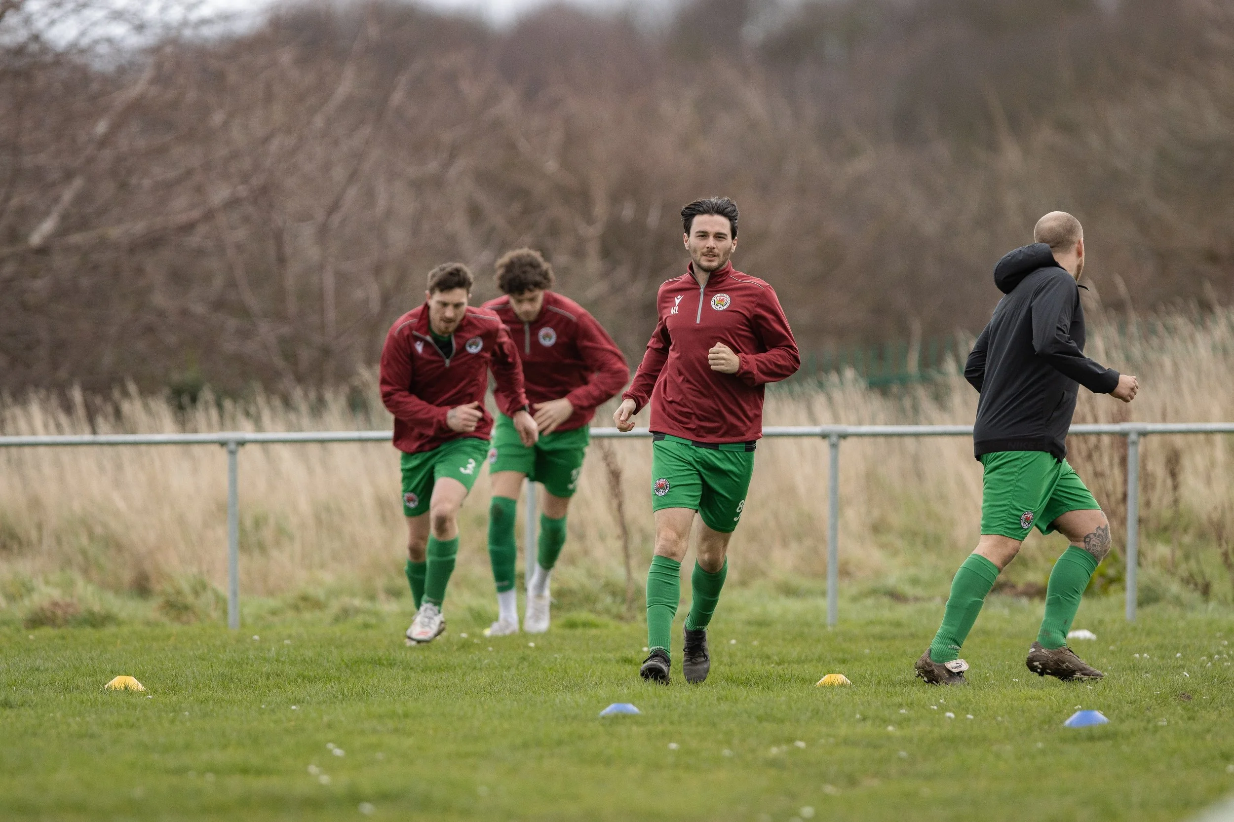 A group of soccer players training outdoors on a grassy field with training cones, wearing red jackets and green shorts, with one person running and others jogging.