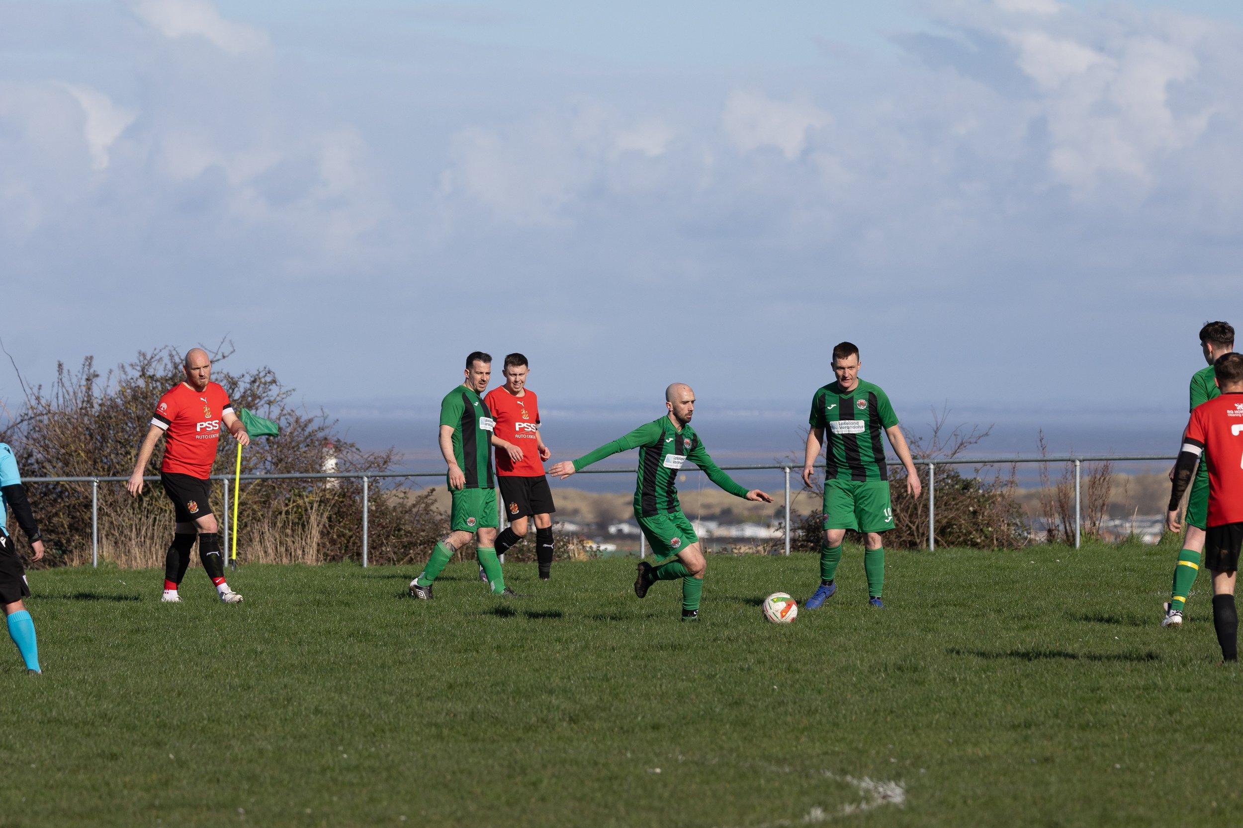 Soccer players on a grassy field during a match, wearing green and red jerseys, with some players standing and one preparing to kick the ball, under a partly cloudy sky.