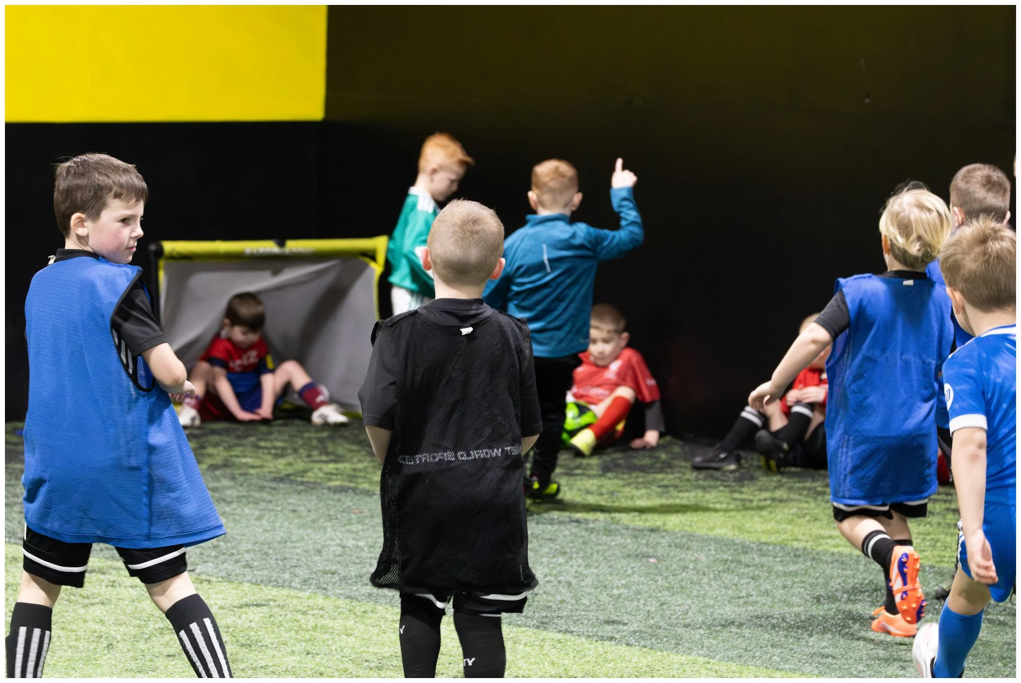 Young boys playing indoor soccer, some wearing blue jerseys, with a few sitting on the ground in red jerseys, near a small goal on a green turf field.