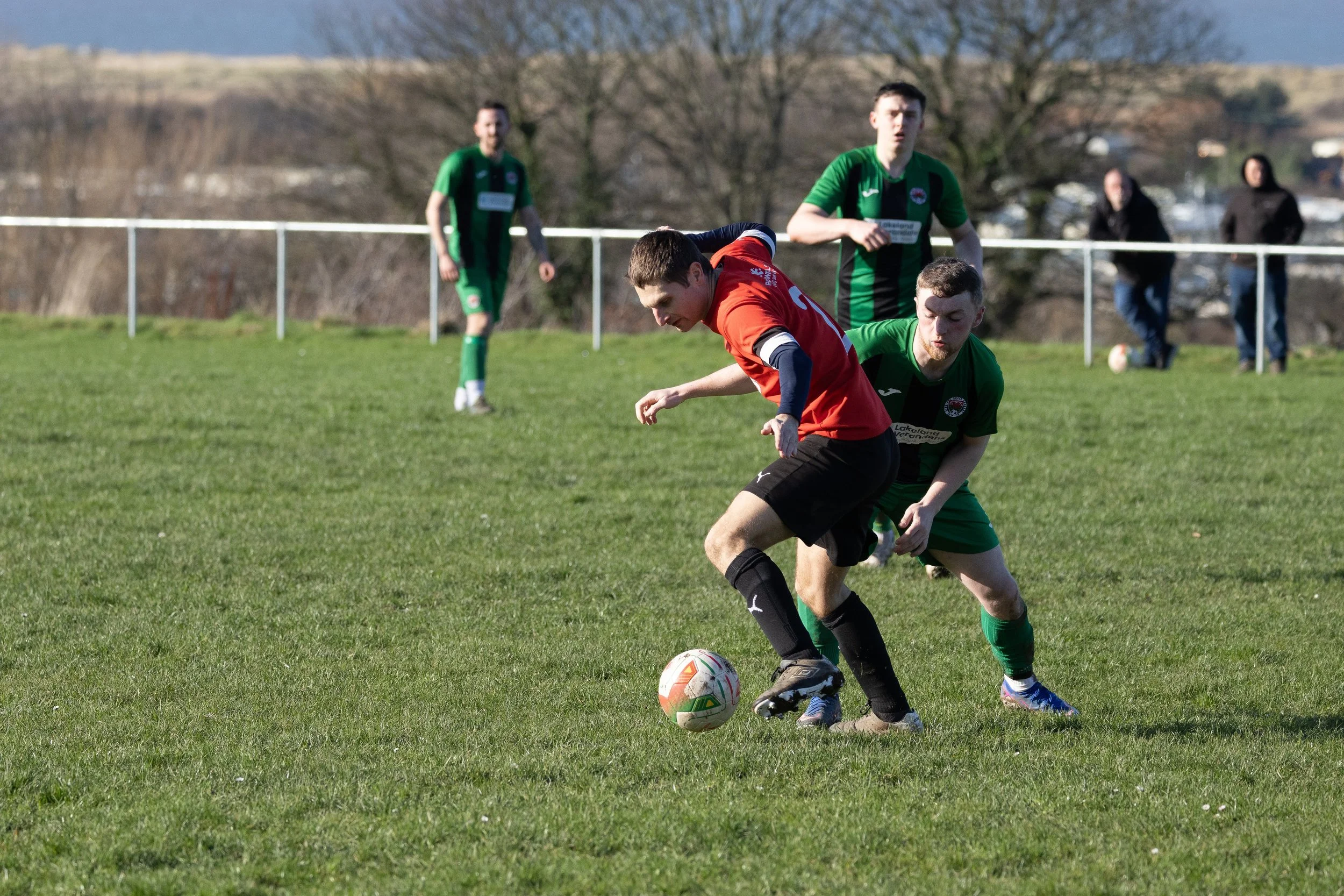 Soccer players compete for the ball on a grassy field, with three players in green jerseys and one on red jersey in foreground, and spectator in background.