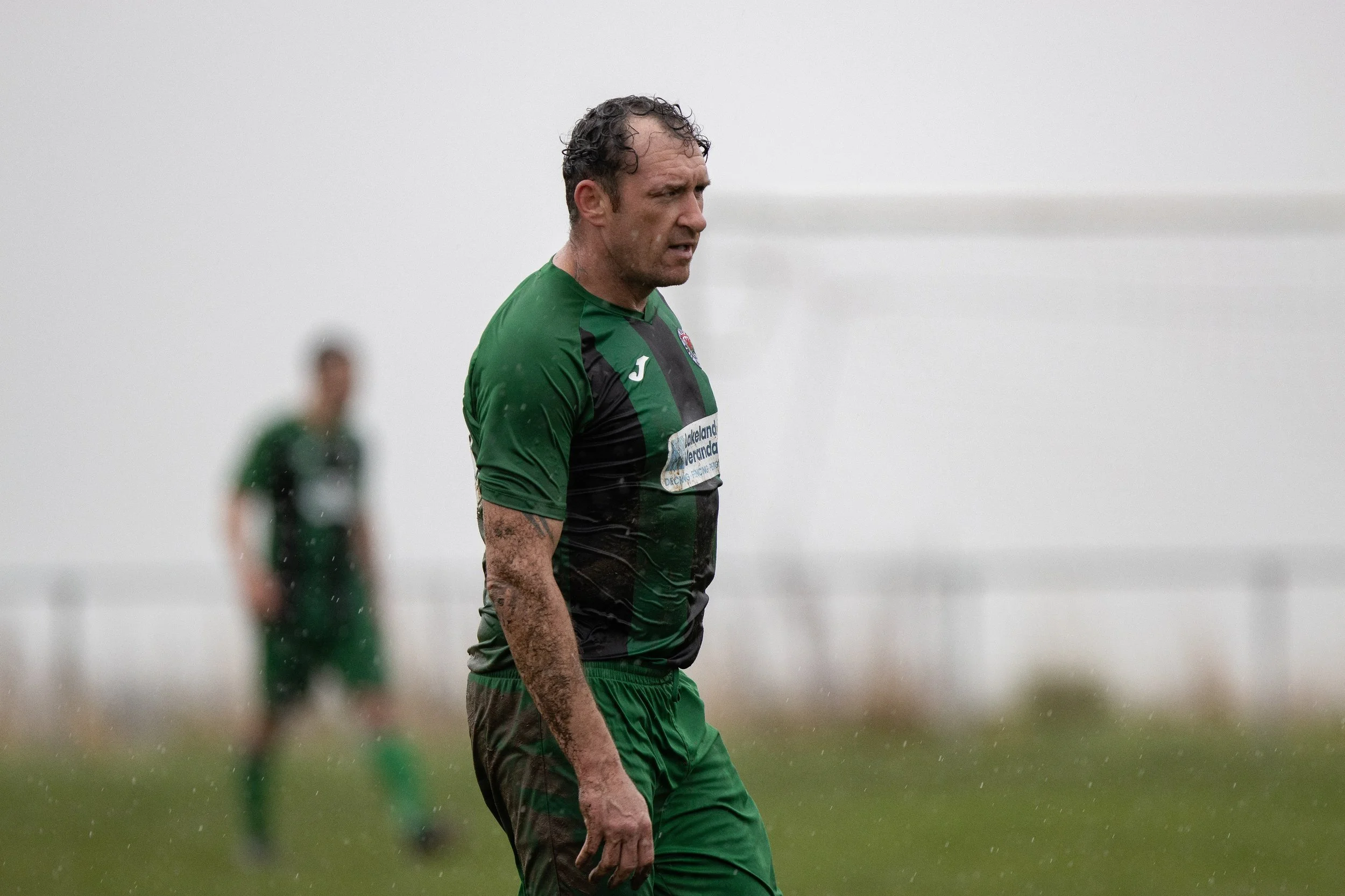 A shirtless male athlete with muddy arms and wet hair, wearing a green and black sports jersey, standing on a rainy soccer field with a blurred background of another player and a goalpost.
