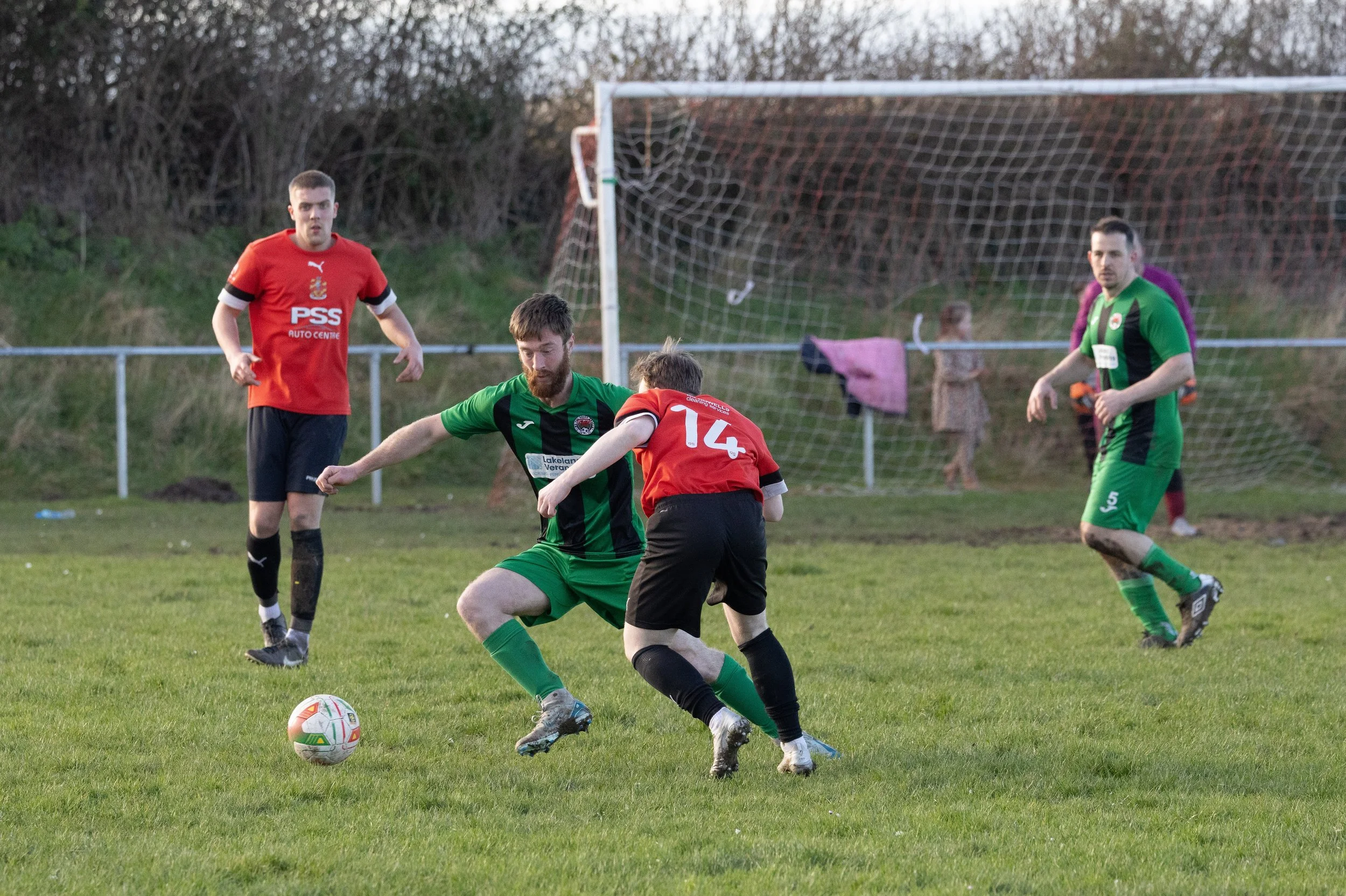 A soccer game with players in red and green uniforms on a grassy field. One player in green is kicking the ball while a player in red tries to block. Two other players in green and red are in the background.