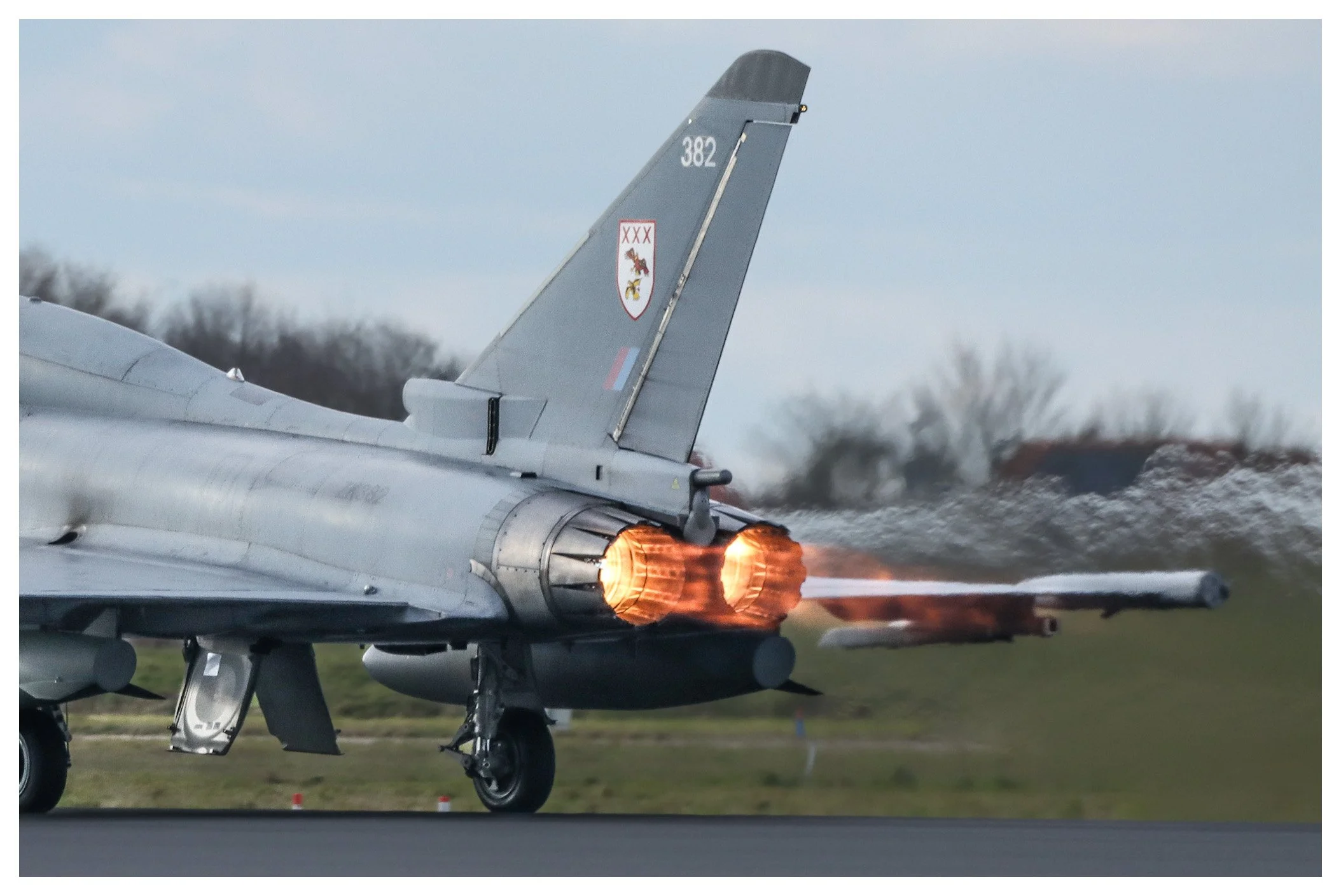 Military jet aircraft taking off with afterburners engaged, showing flames and heat distortion, on a runway with a cloudy sky and trees in the background.