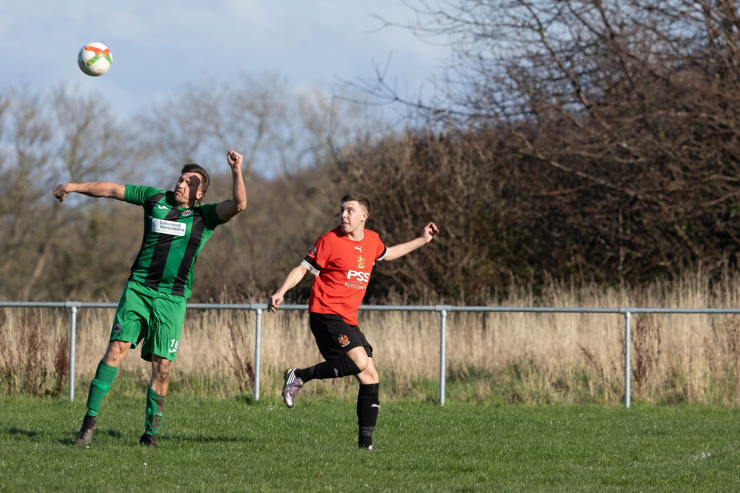 Two soccer players, one in a green and black uniform and the other in a red jersey and black shorts, are on a grassy field with a metal fence and trees in the background. The player in green is heading the ball, while the player in red is running nea