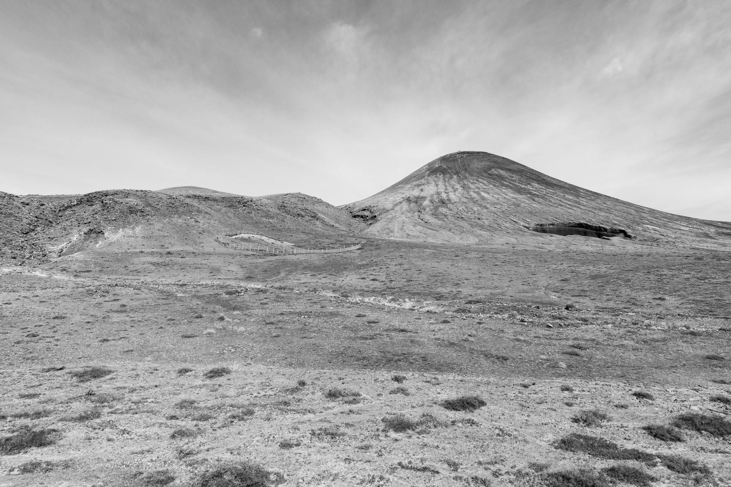 A black and white landscape featuring a large, gently sloping mountain in the background with a flat, arid terrain in the foreground. The sky is cloudy and overcast.