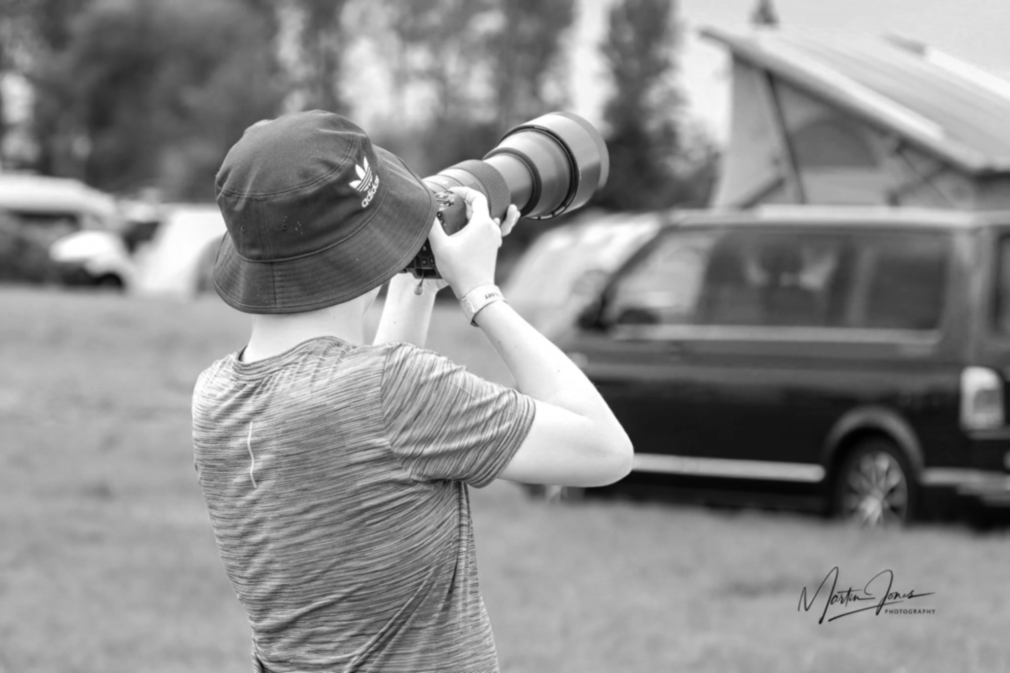 A person wearing a bucket hat and striped t-shirt is taking a photograph with a camera featuring a large telephoto lens outdoors, with cars parked in the background.