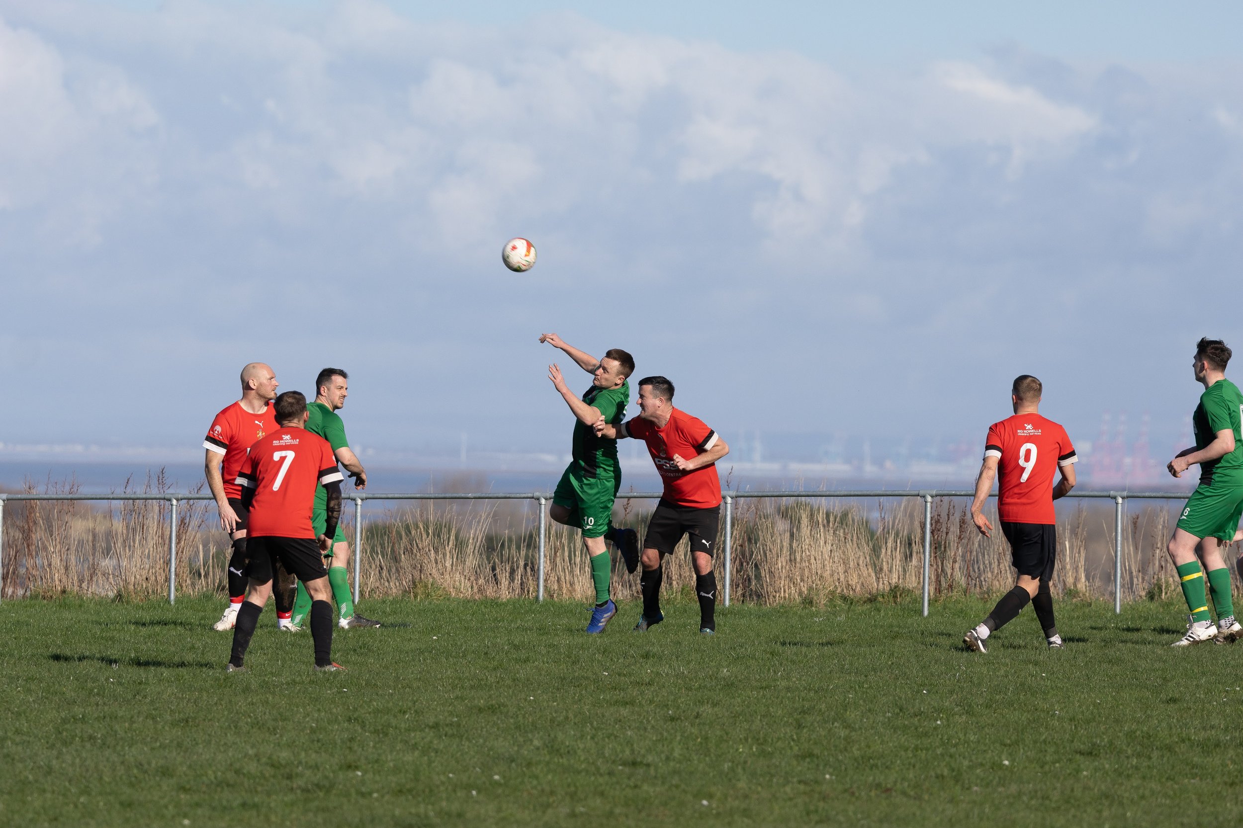 Soccer players in a match, with two players jumping for the ball and others watching, on a grassy field under a partly cloudy sky.