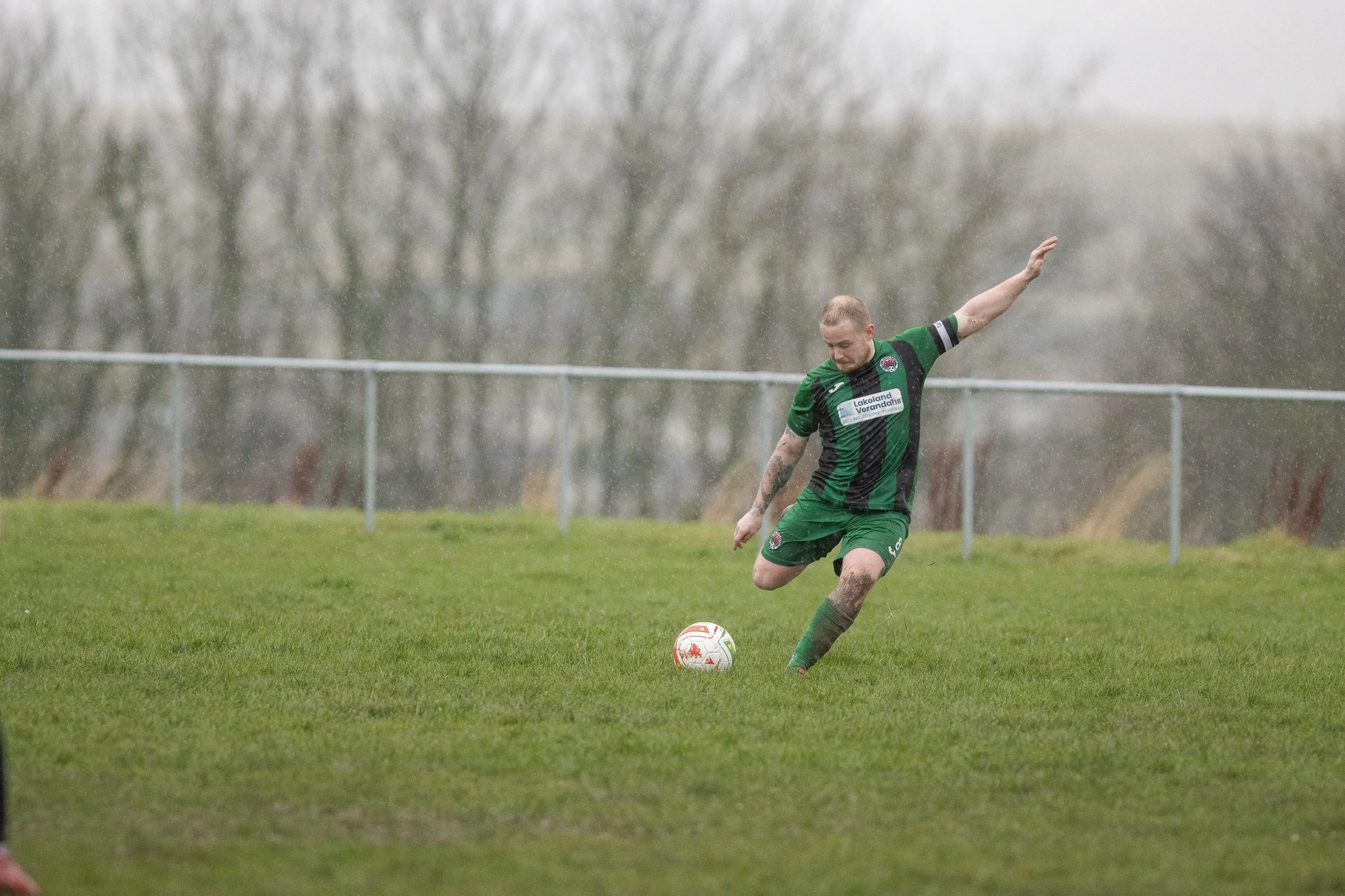 A soccer player in a green and black uniform kicking a ball during rain on a grassy field.