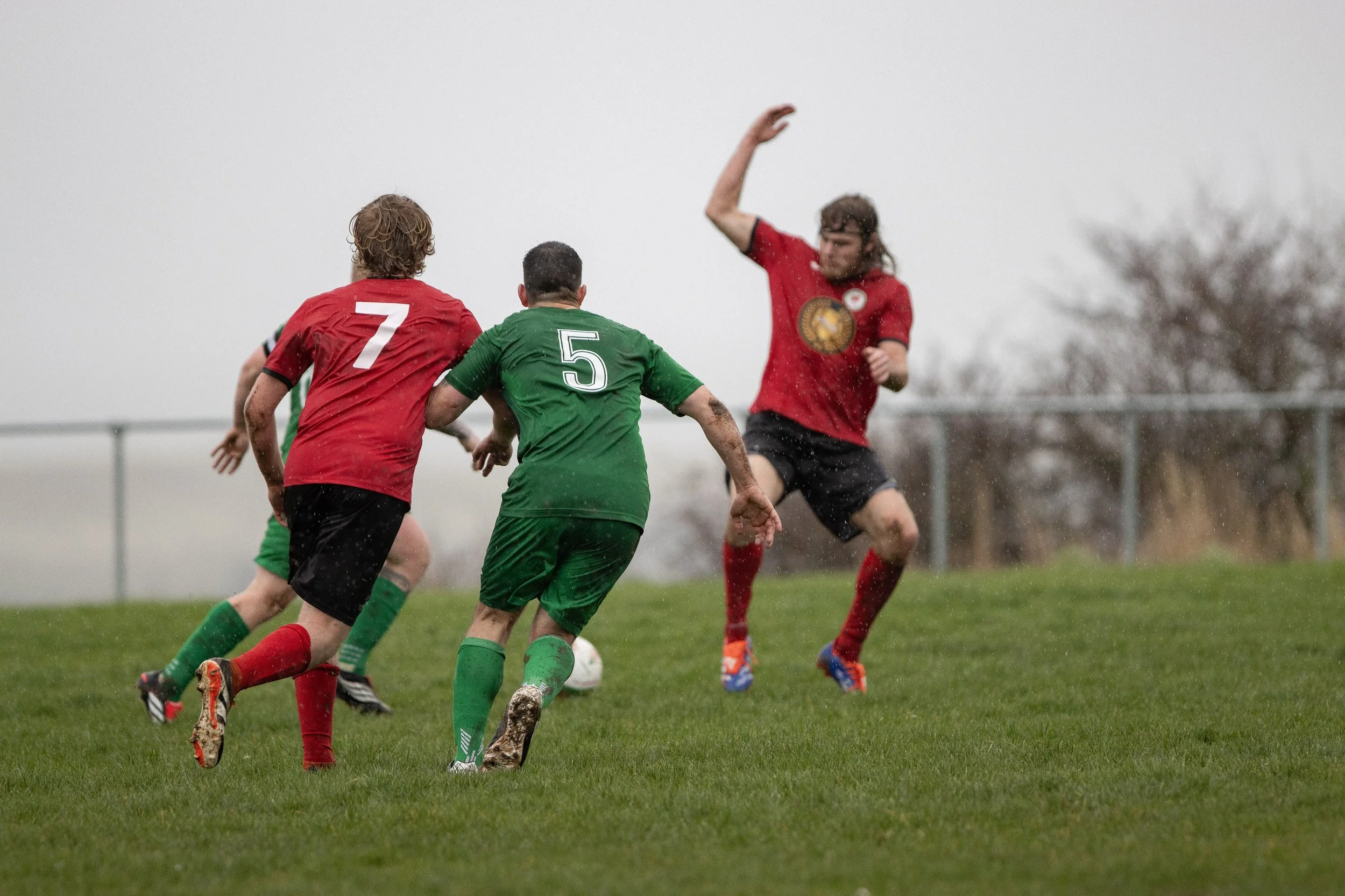 Four soccer players on a rainy field, two in red jerseys and two in green, with one player in motion near the goal.