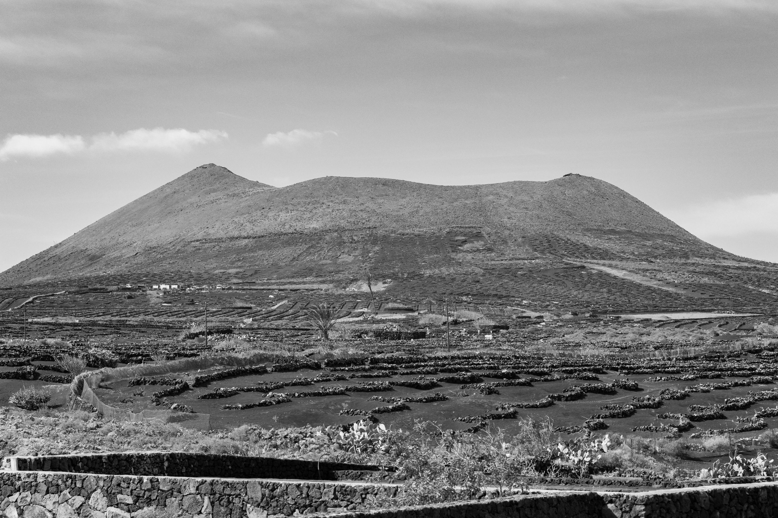 Black and white photo of a twin-peaked volcanic mountain with terraced fields and farm structures at the base.
