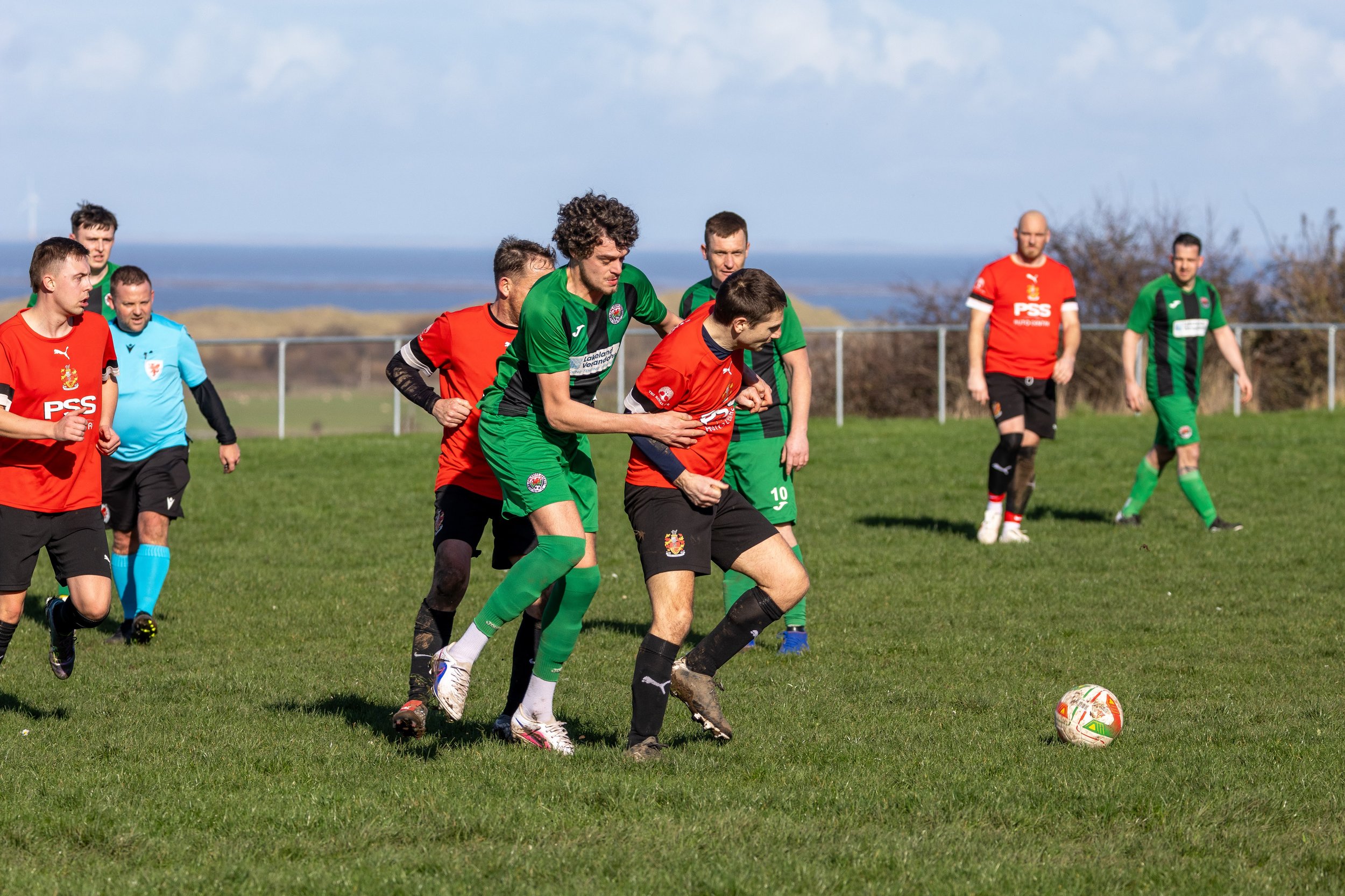Soccer players in red and green uniforms contest for the ball on a grassy field, with some players running and others watching under a partly cloudy sky.