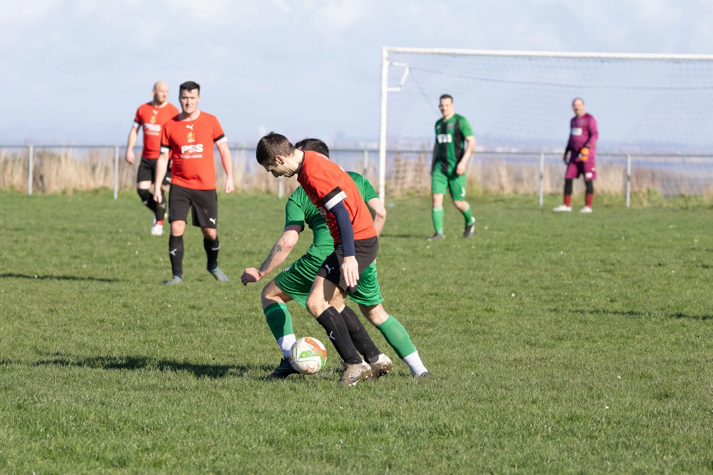 Two soccer players in green and red uniforms competing for the ball on a grass field, with four other players and a goal in the background.