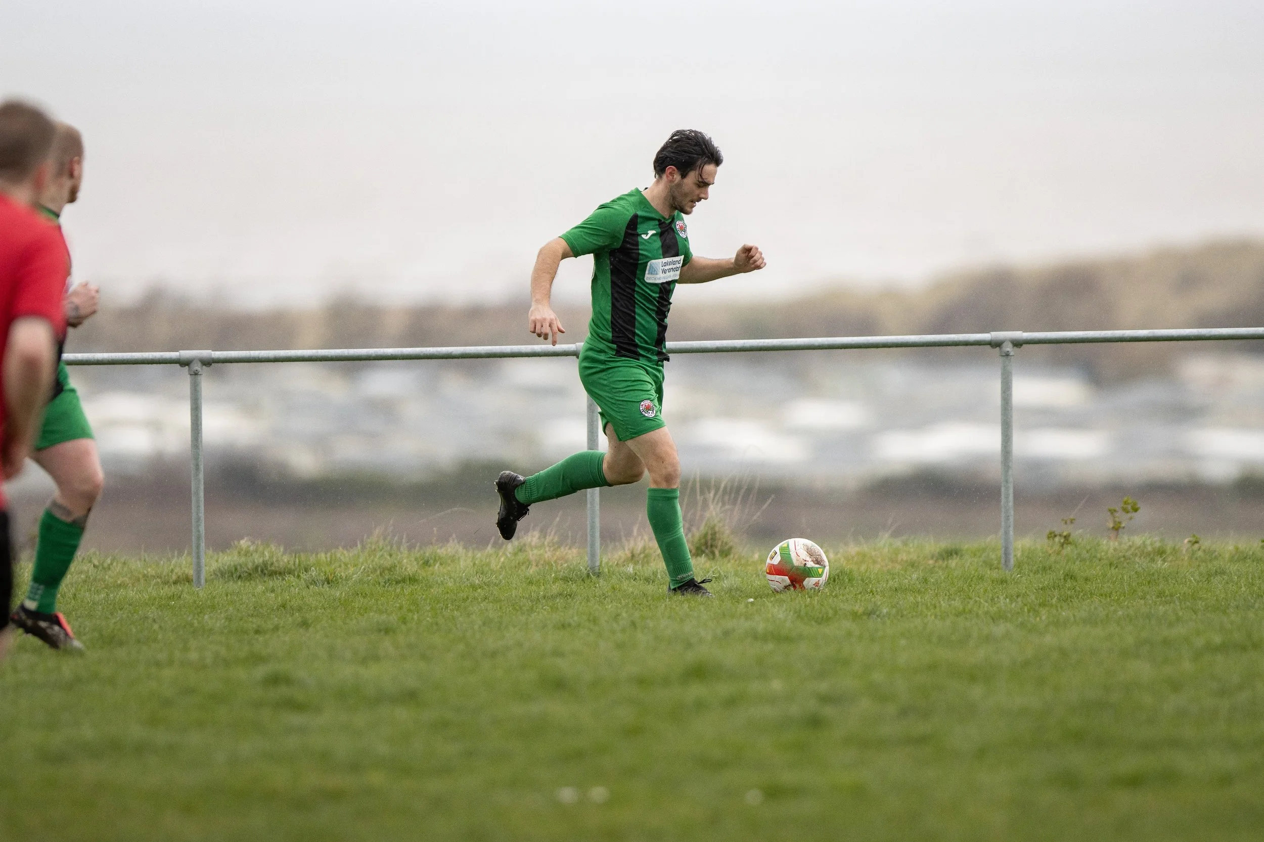 A soccer player in a green and black uniform kicking a ball on a grassy field during a game.