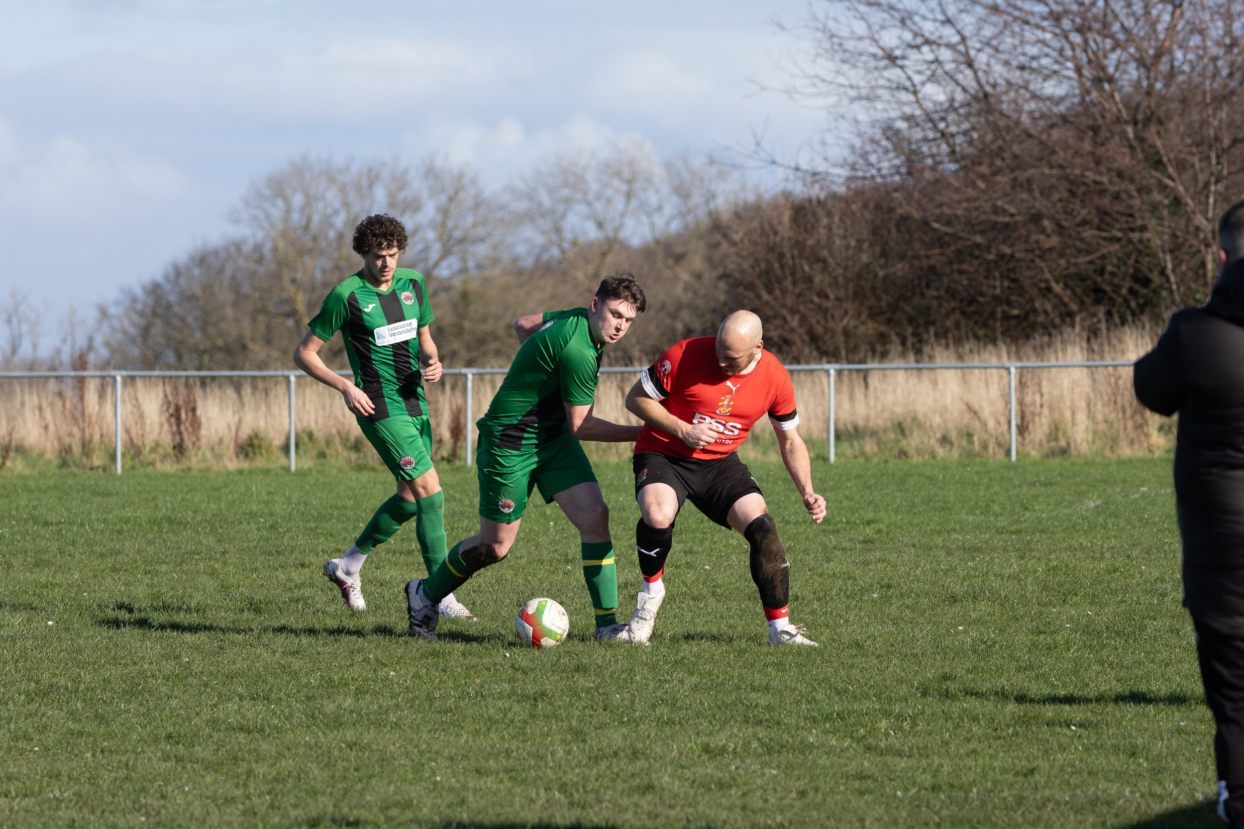 Three soccer players on a grassy field competing for the ball while a person watches on the right.