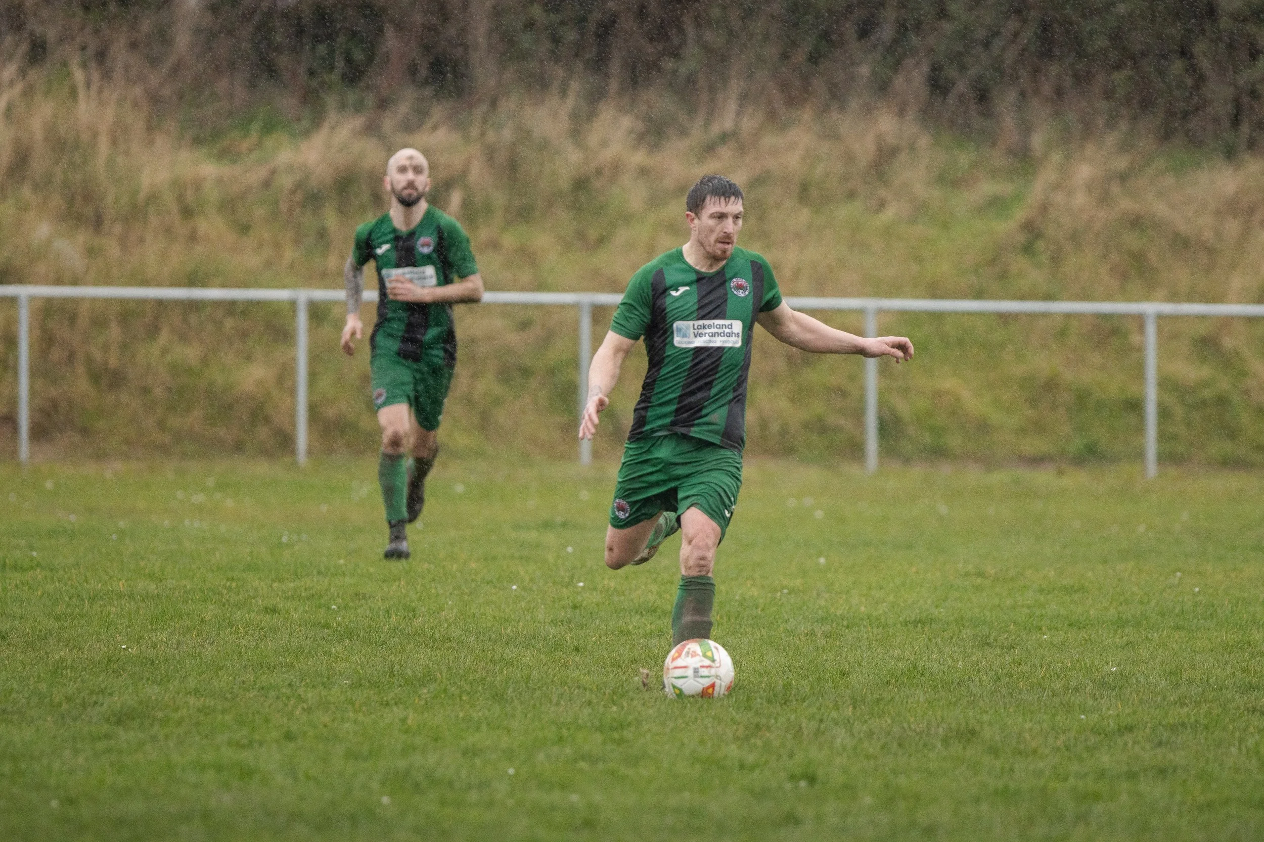 Two soccer players in green and black uniforms on a rain-soaked field, with one player dribbling the ball and the other running in the background, surrounded by a muddy strip on their jerseys.