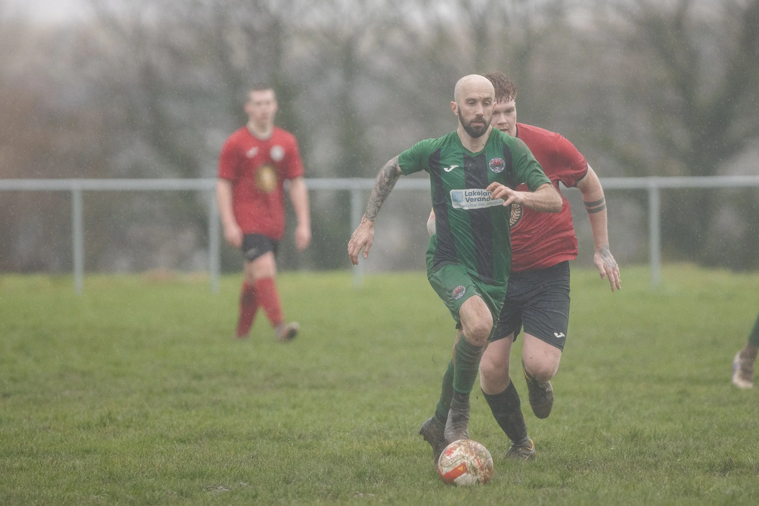 Two soccer players, one in a green and black uniform and the other in a red and black uniform, racing for the ball on a rainy day during a match.